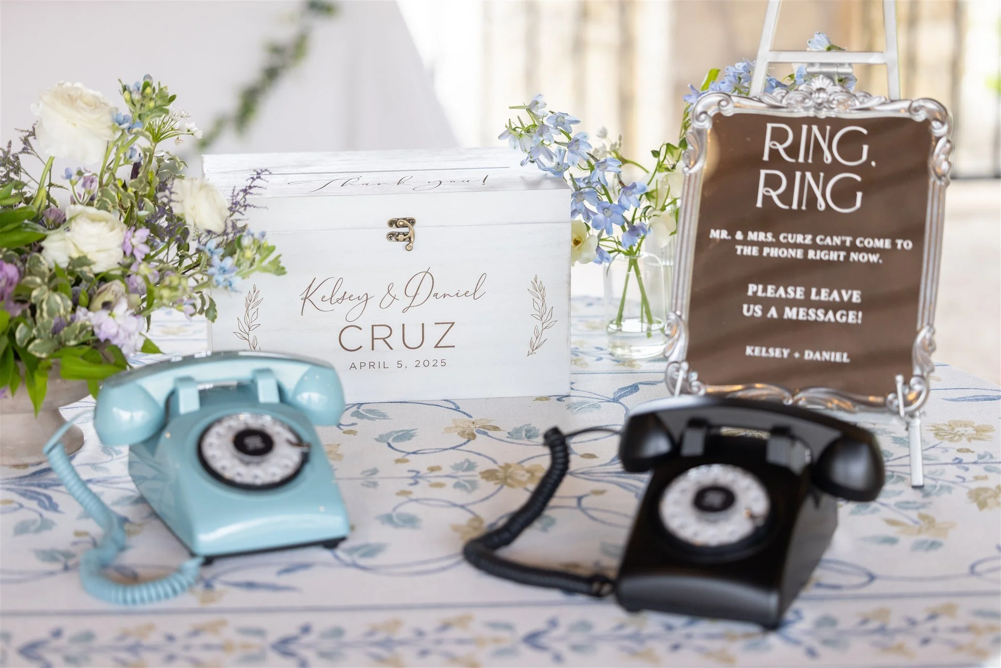 A wedding reception table with a white wooden box, floral arrangements, a sign about leaving messages, and two vintage rotary phones, one light blue and one black.