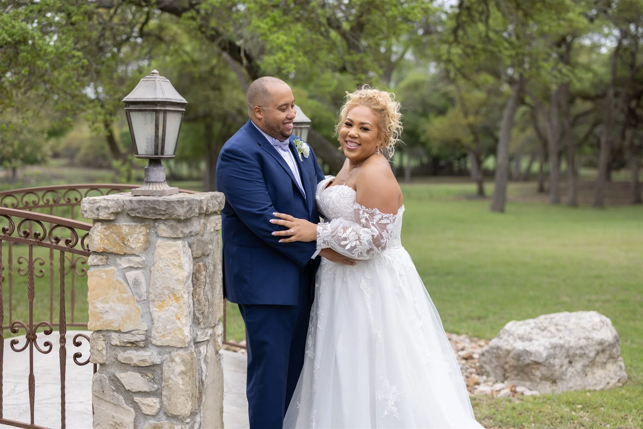 A bride and groom in wedding attire smiling and embracing outdoors in a park-like setting with trees and grass.