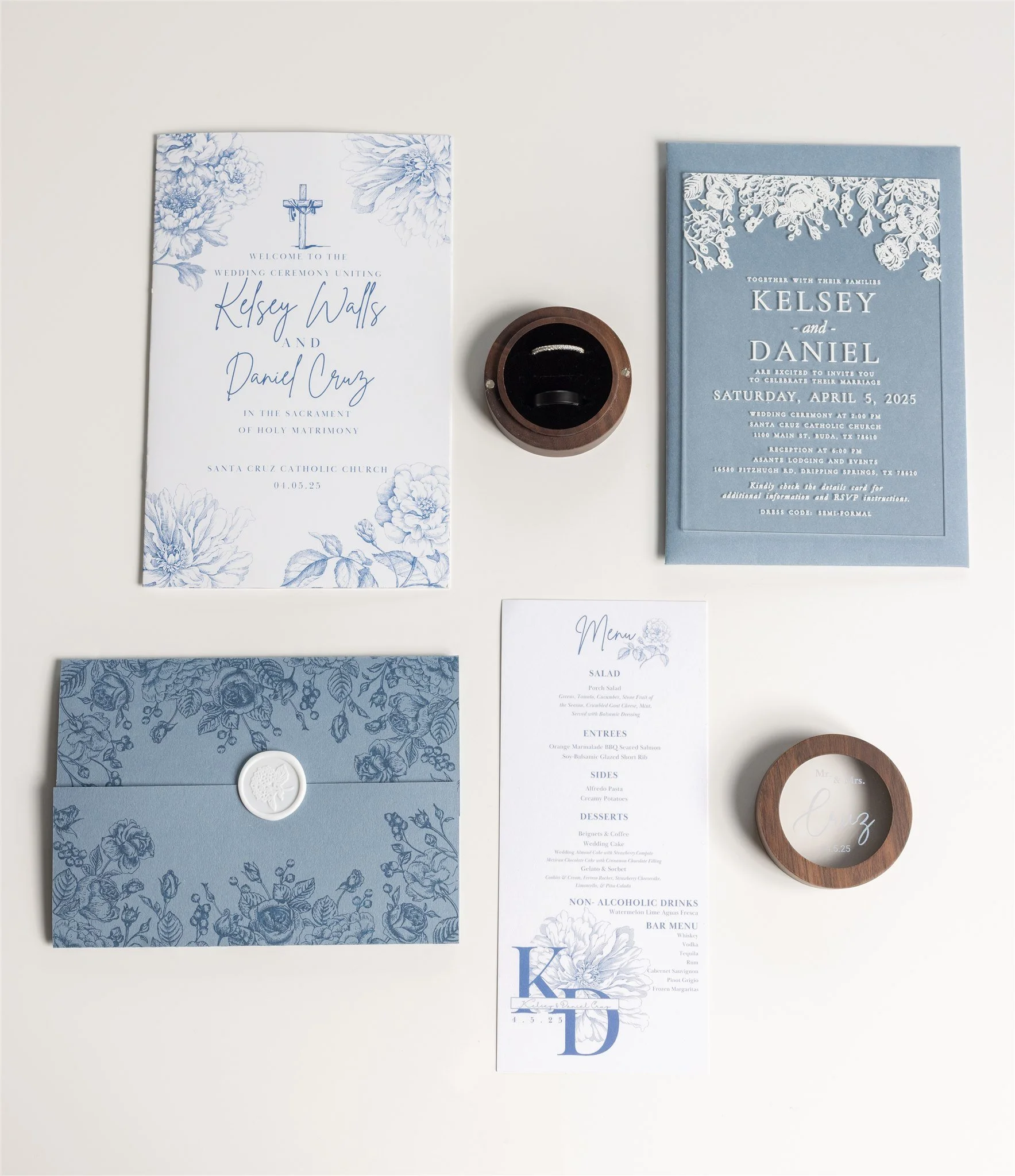 Wedding invitation suite with blue and white floral designs, a menu card, a blue envelope with a wax seal, and a round glass-top wooden ring box on a white background.