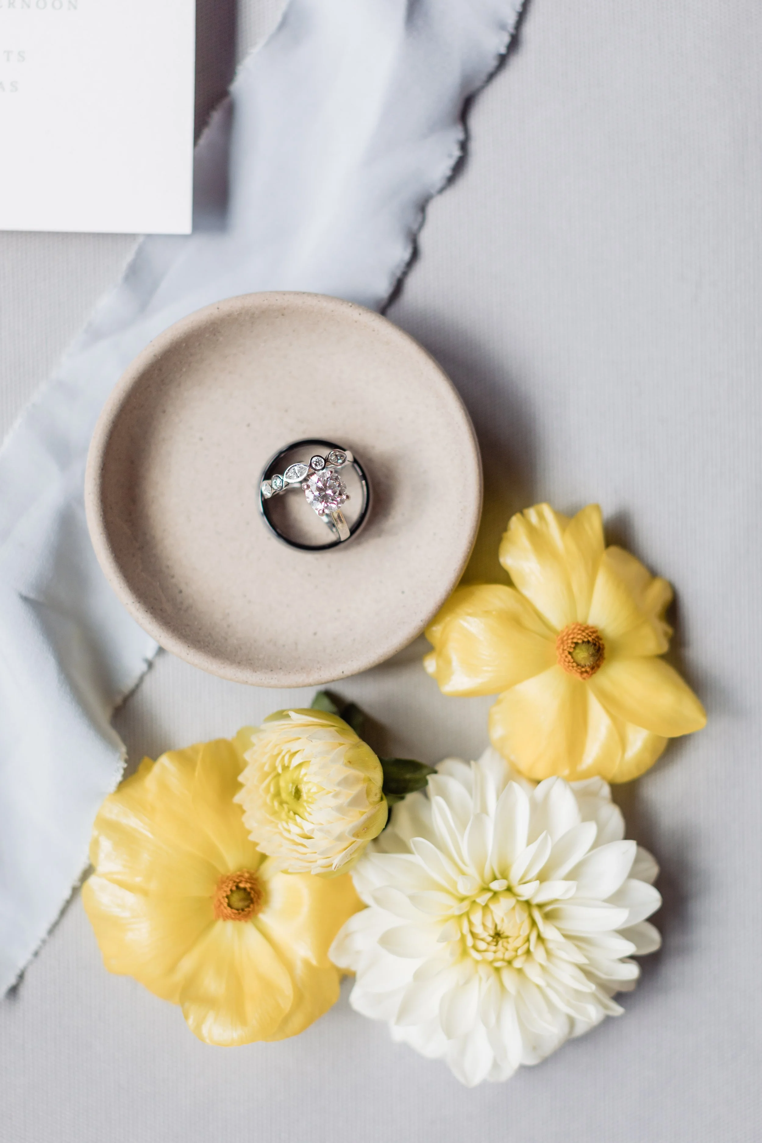 A wedding ring with a pink gemstone and diamonds in a beige bowl, surrounded by yellow and white flowers on a white cloth surface.