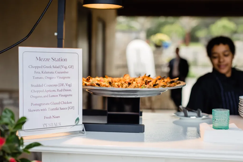 Plate of assorted small plates including Greek salad, couscous, and chicken skewers on a serving stand at a buffet counter.