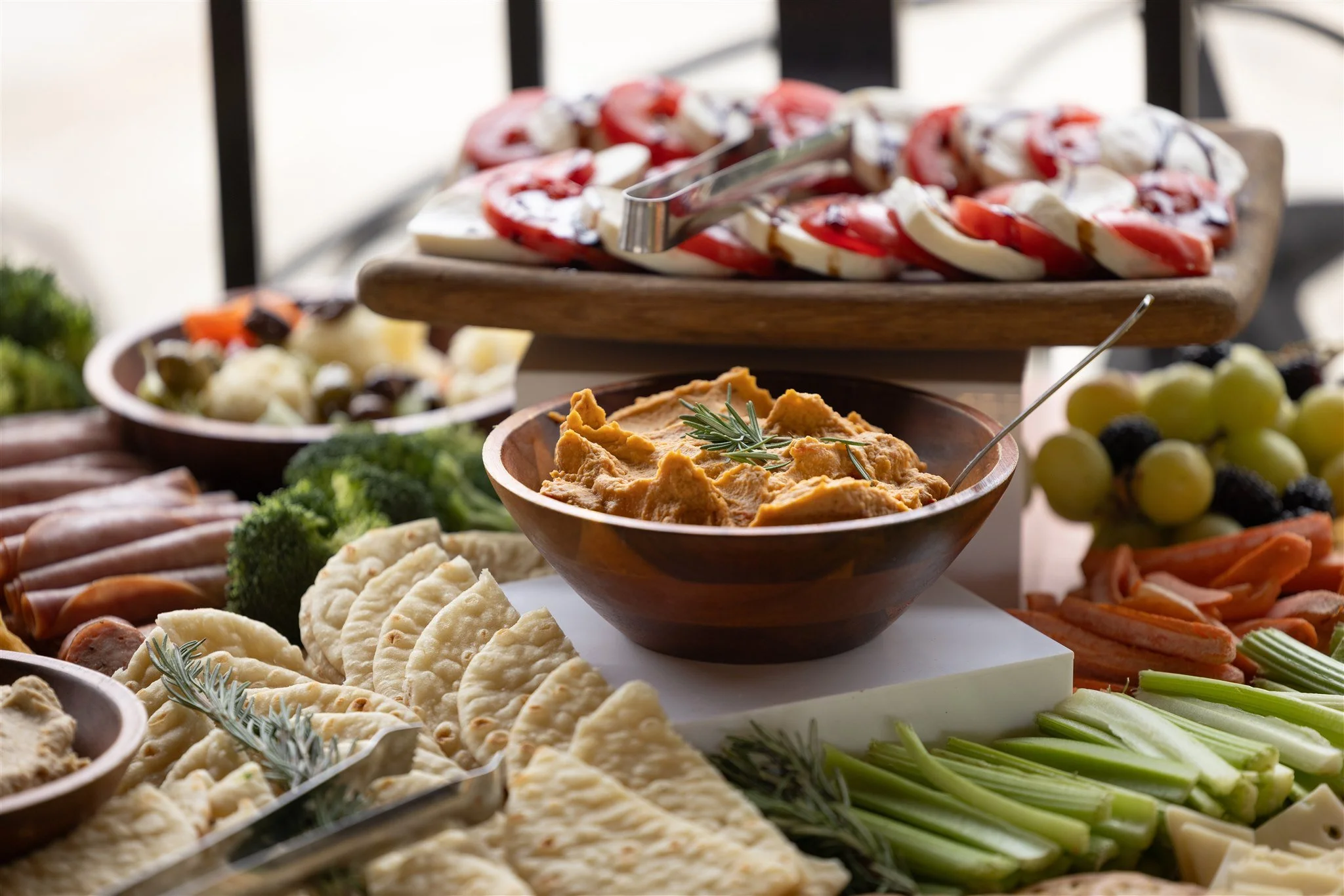 A variety of food including hummus, grapes, celery, cheese, meat, bread, and topped with tomato and mozzarella salad on a wooden platter