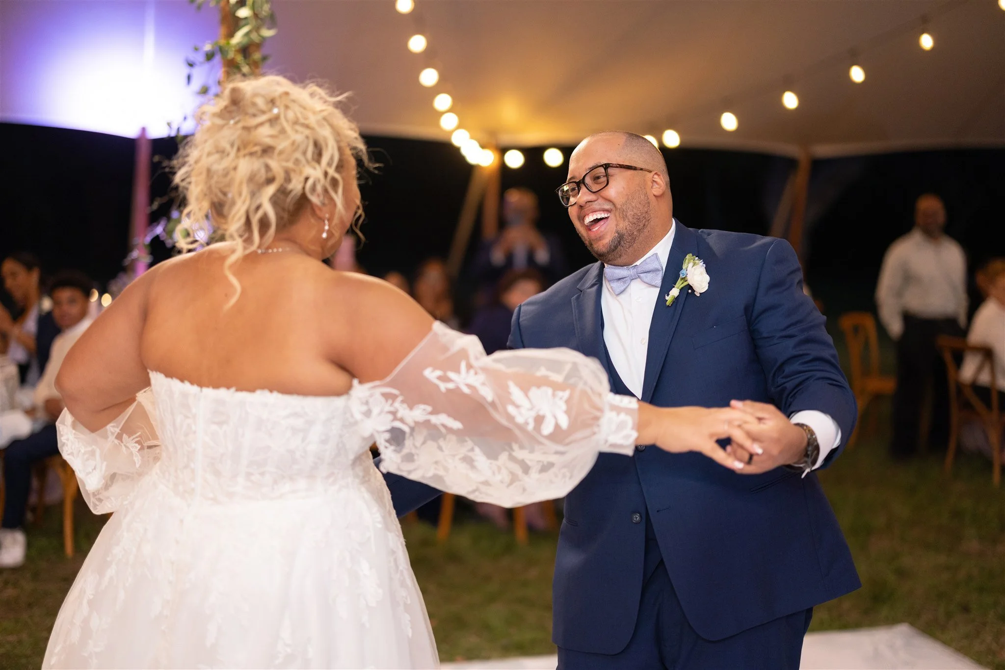 A bride and groom dancing at their wedding reception, with string lights hanging overhead and guests seated in the background.