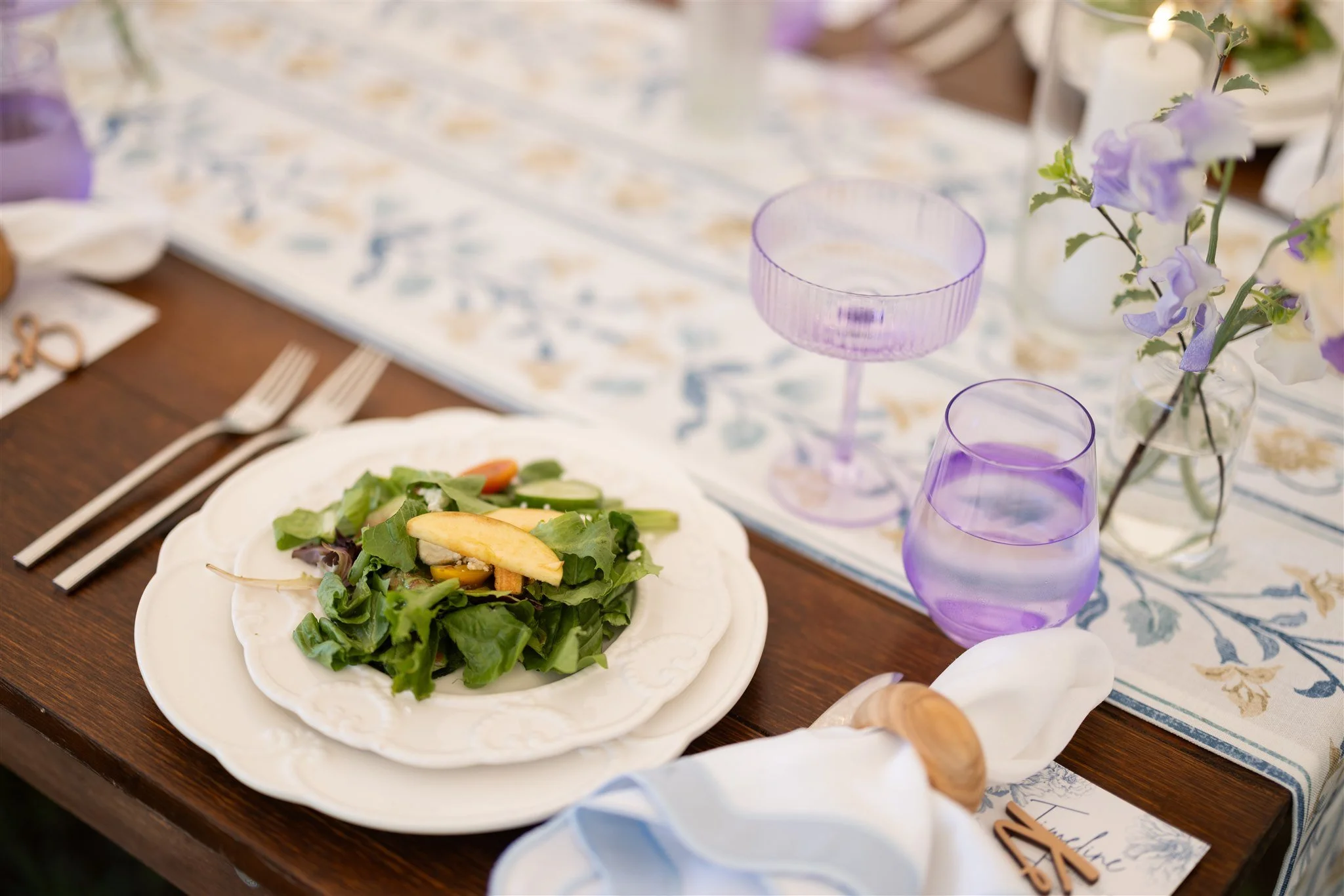 A formal dining table with a white plate containing a salad with greens and apple slices, two forks, a lavender glass, a lavender goblet, a small glass vase with purple and white flowers, a white napkin, and a wooden pepper shaker.