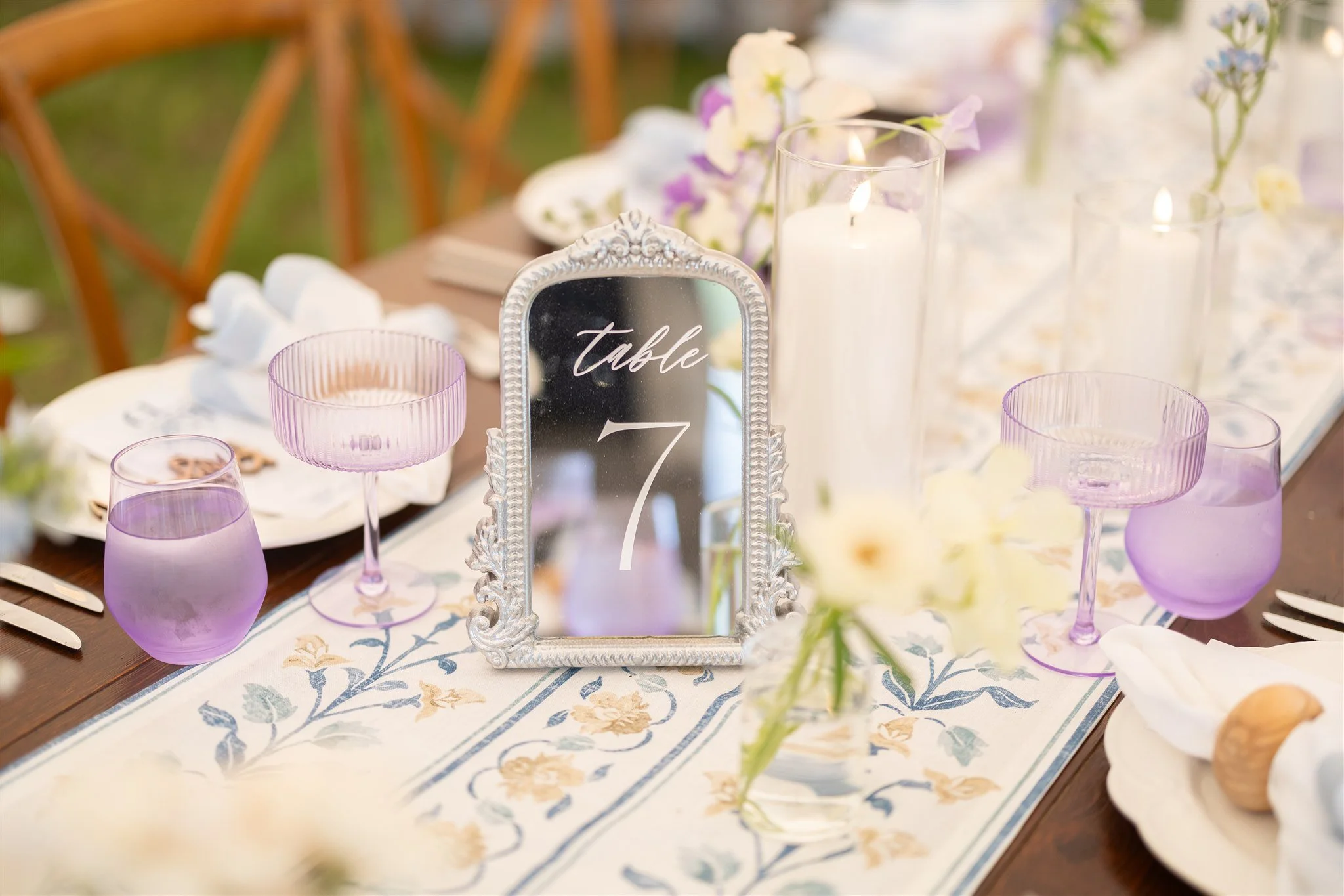 Decorated dining table with a table number mirror, purple glasses, candles, flowers, and elegant tableware.