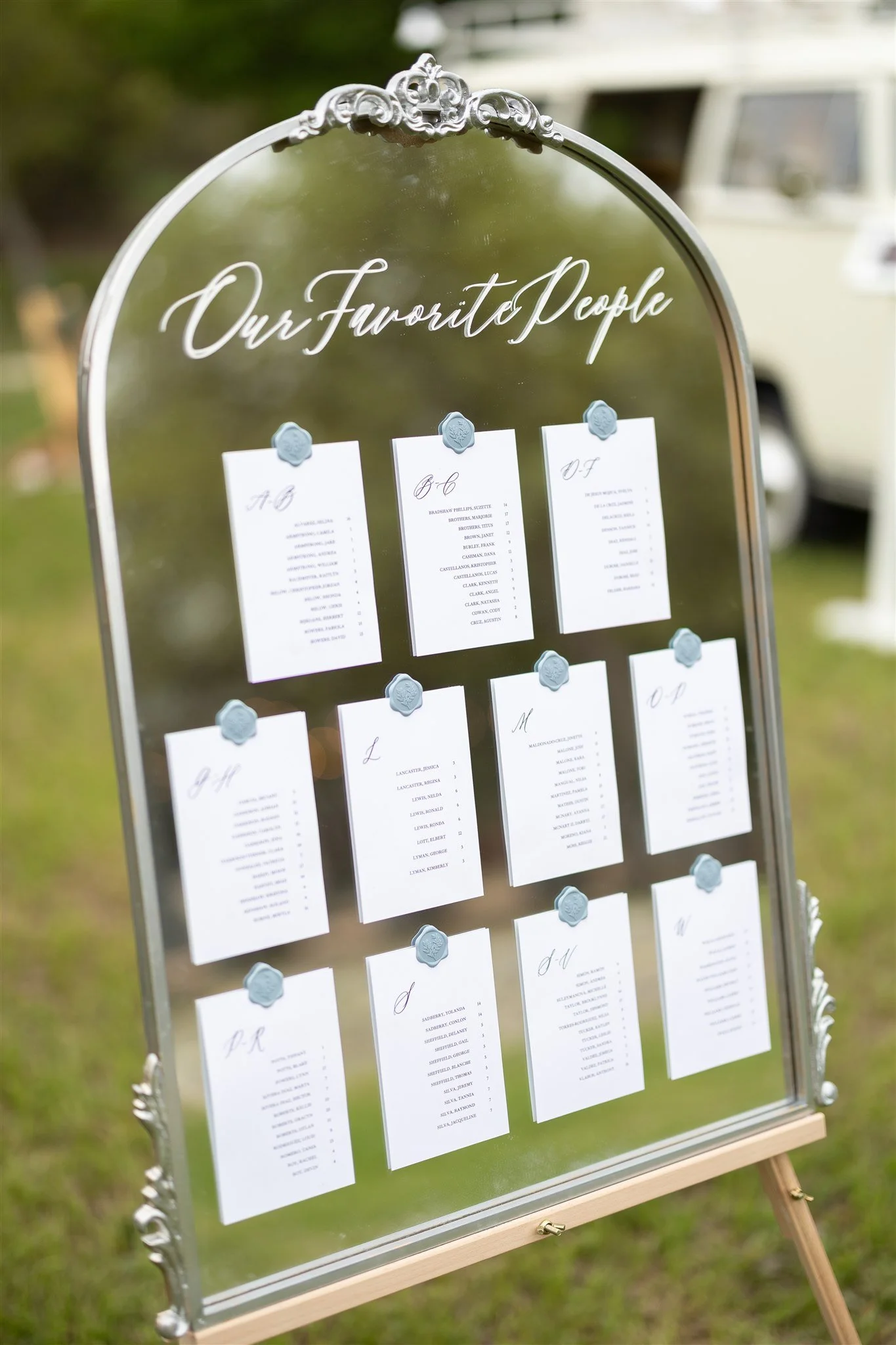 Mirror seating chart display titled "Our Favorite People" with table arrangements on white cards, held by blue wax seals, outdoors on a grassy area.