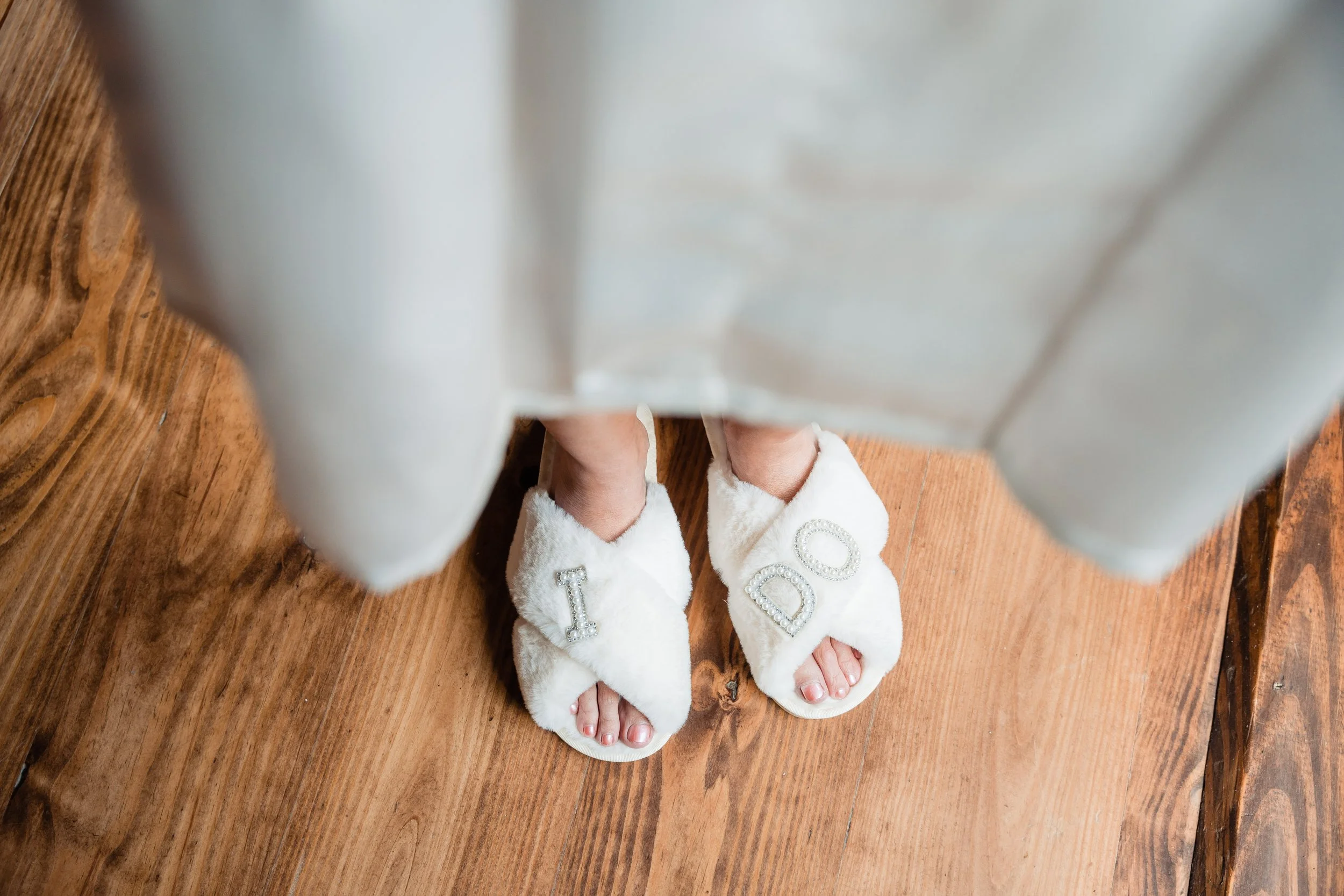 Person wearing white slippers with rhinestone letters spelling DID, standing on a wooden floor.