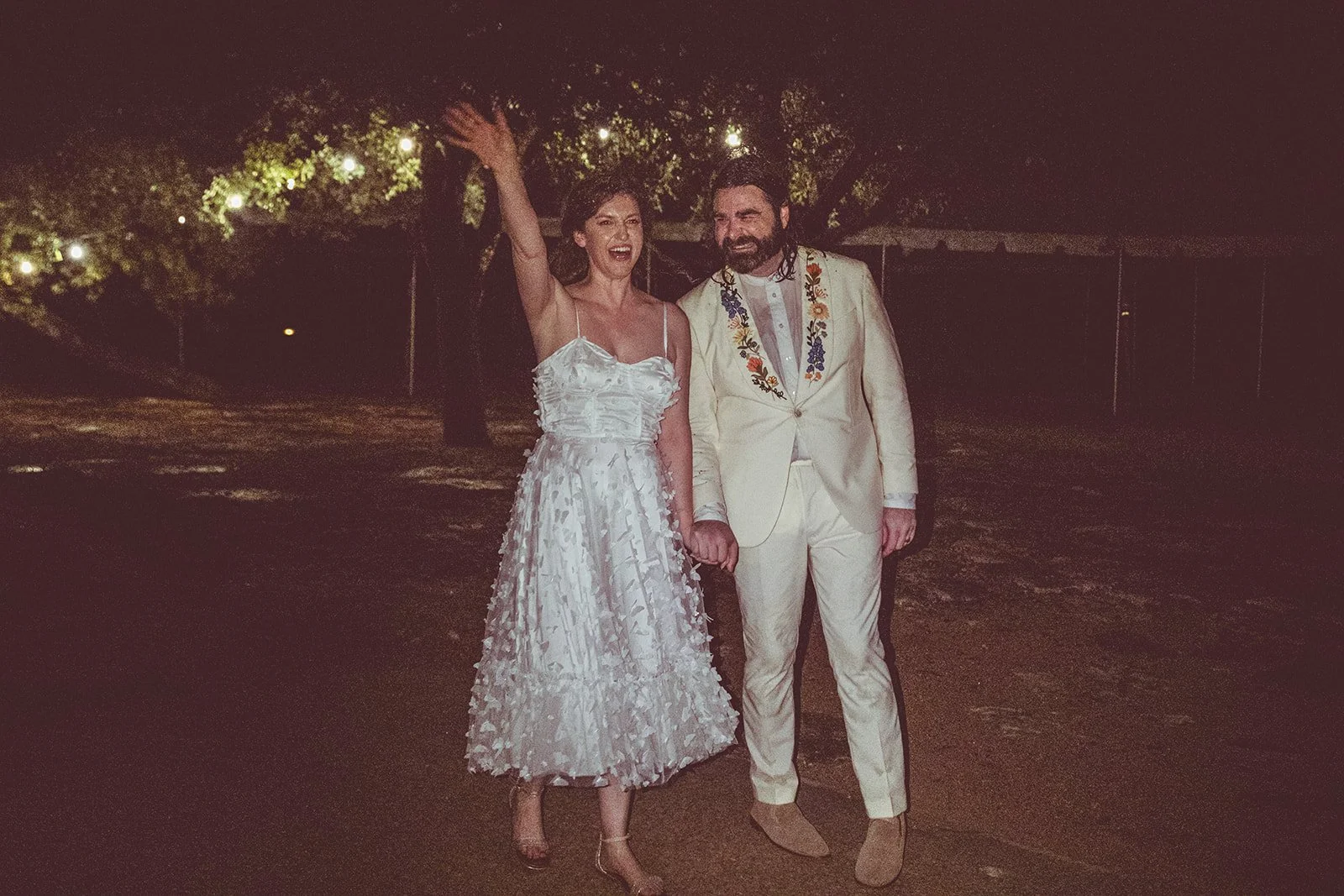 A man and woman dressed in wedding attire, holding hands, standing outdoors at night, with the woman raising her arm and smiling.