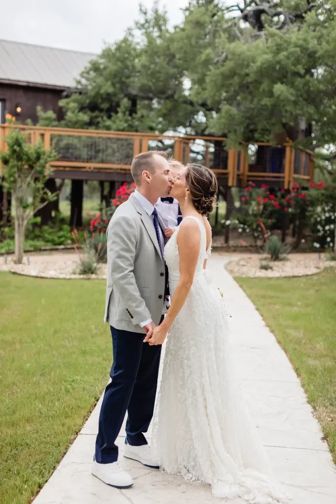 A bride and groom sharing a kiss outdoors on a wedding day, holding hands on a stone pathway amid green grass and garden flowers, with a wooden deck and house in the background.