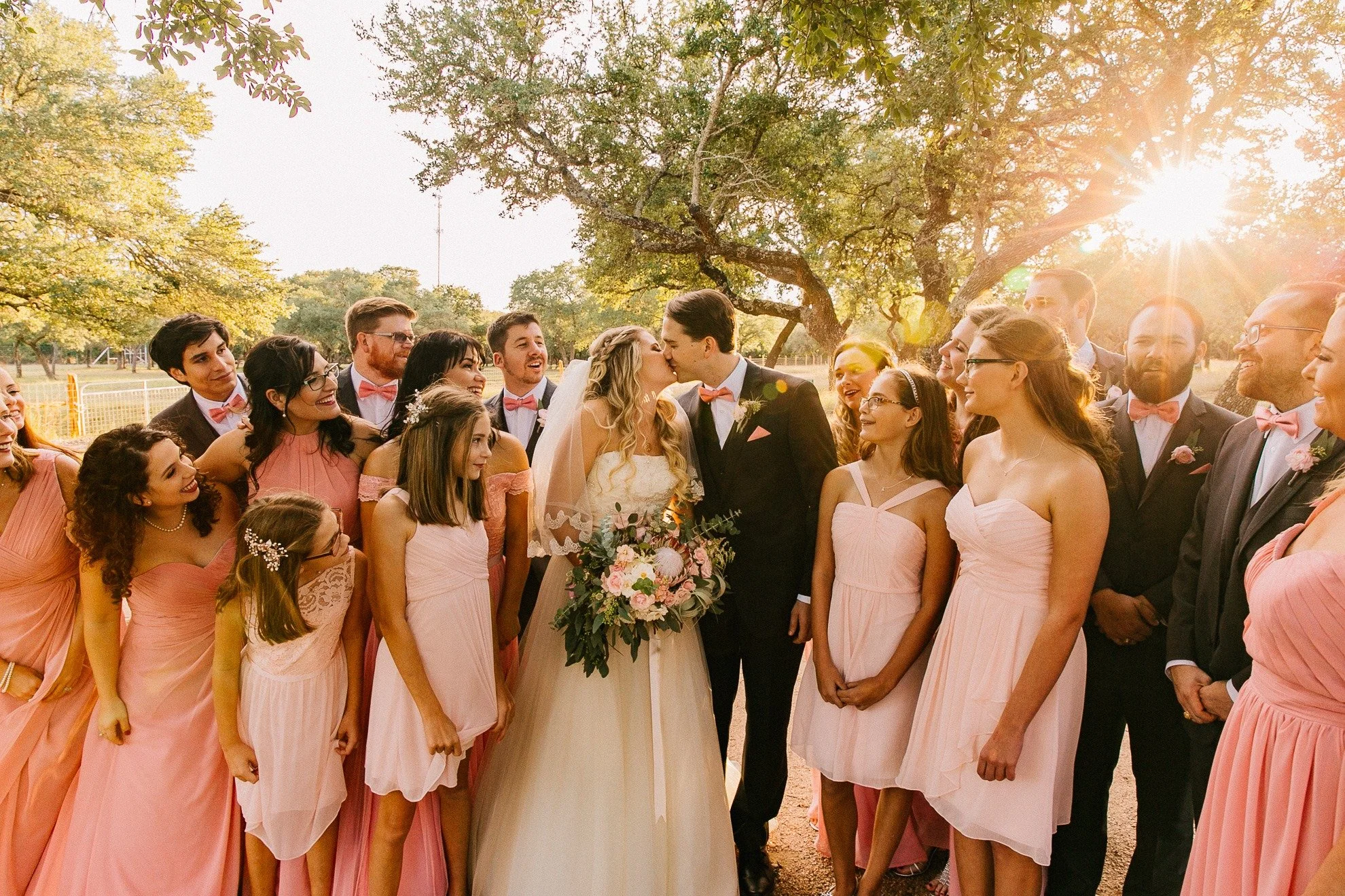 A bride and groom kissing outdoors at sunset, surrounded by bridesmaids and groomsmen in formal attire.