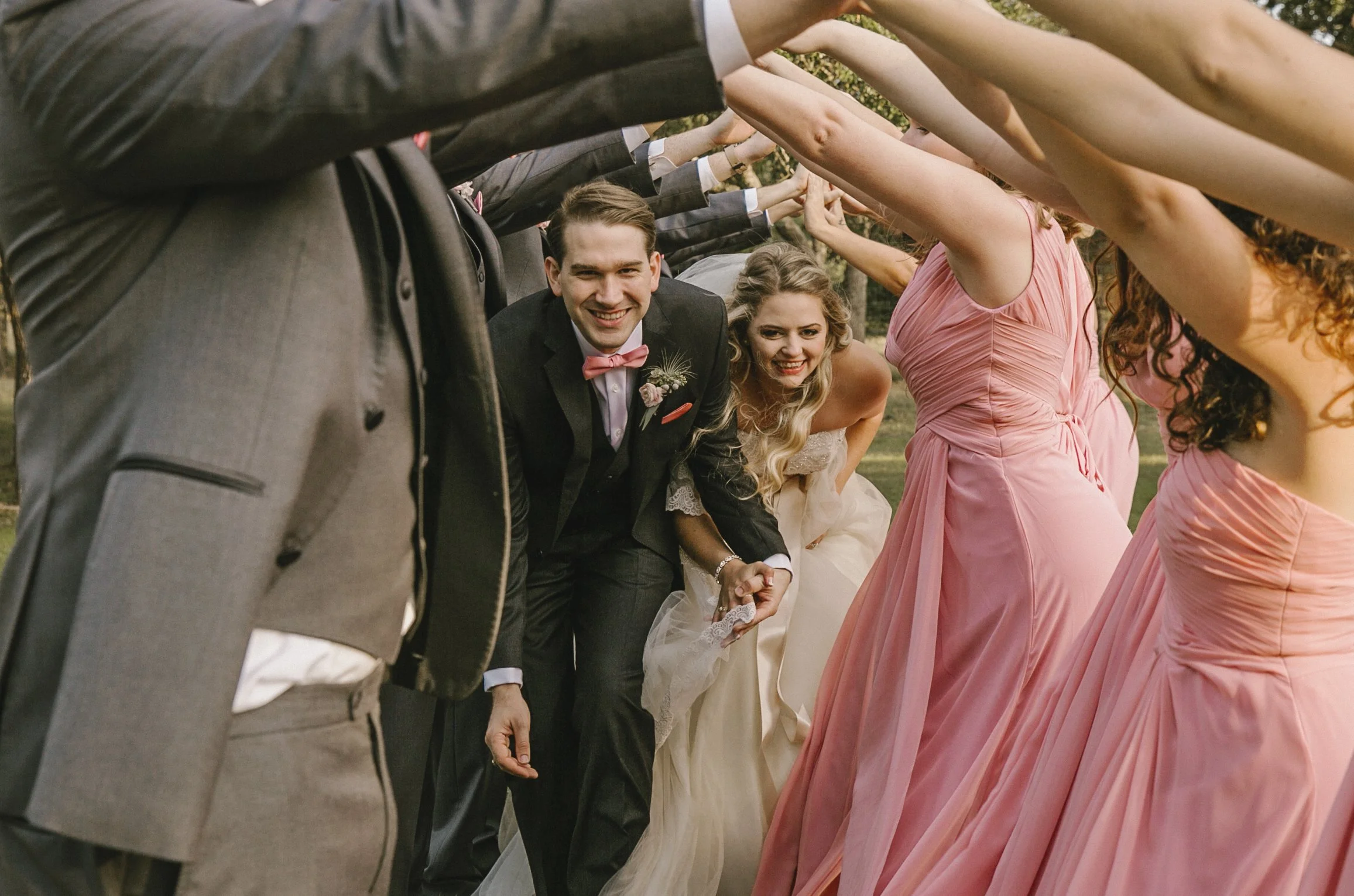 Wedding party with the bride and groom smiling and holding hands, surrounded by bridesmaids and groomsmen forming an arch with their arms.