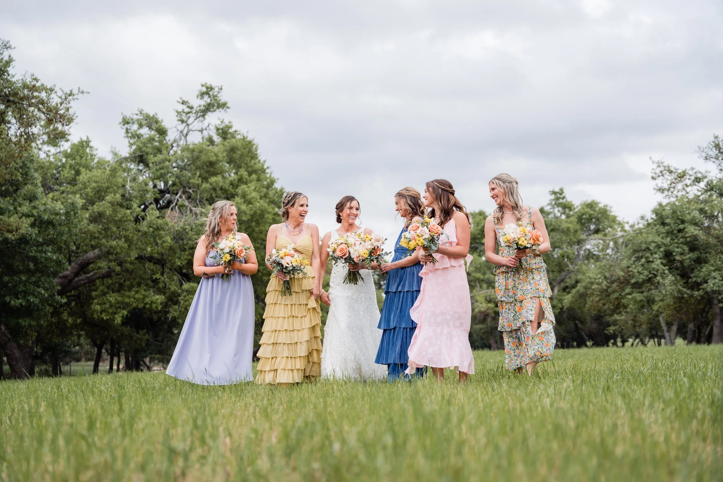 Wedding party of six women in colorful dresses holding bouquets standing on green grass with trees and cloudy sky in the background.