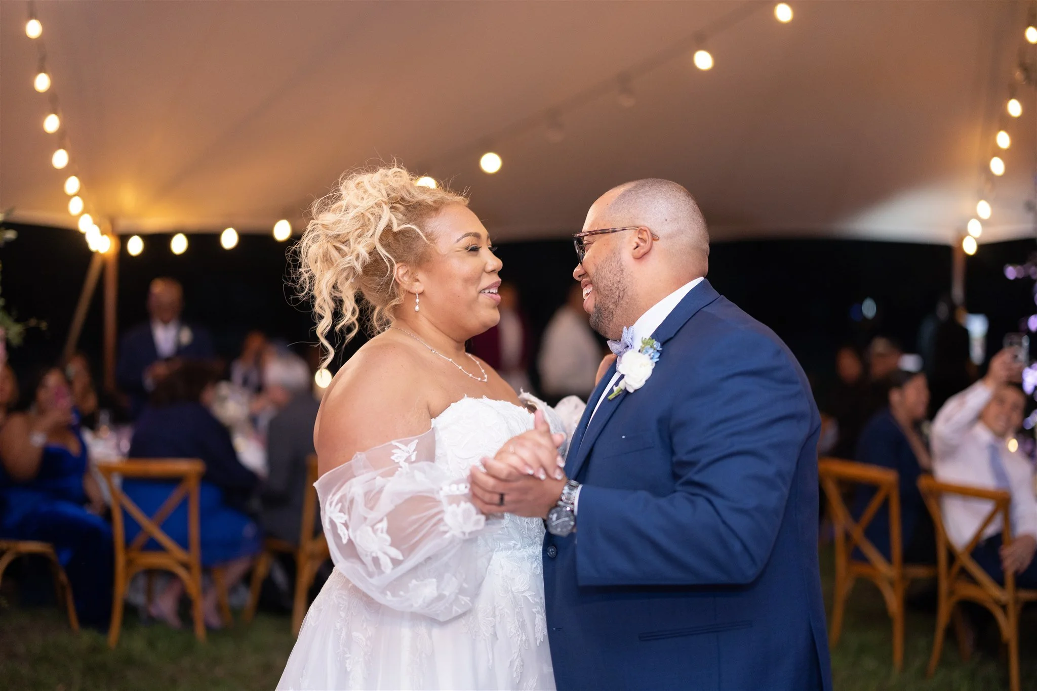 A bride and groom dance at their wedding reception under string lights, smiling warmly at each other, with guests seated in the background.
