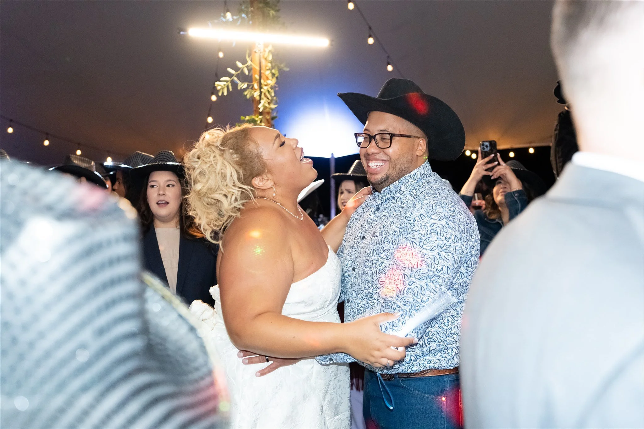 A woman in a white wedding dress and a woman in a blue shirt and black cowboy hat are dancing together at a celebration, surrounded by other people under string lights.