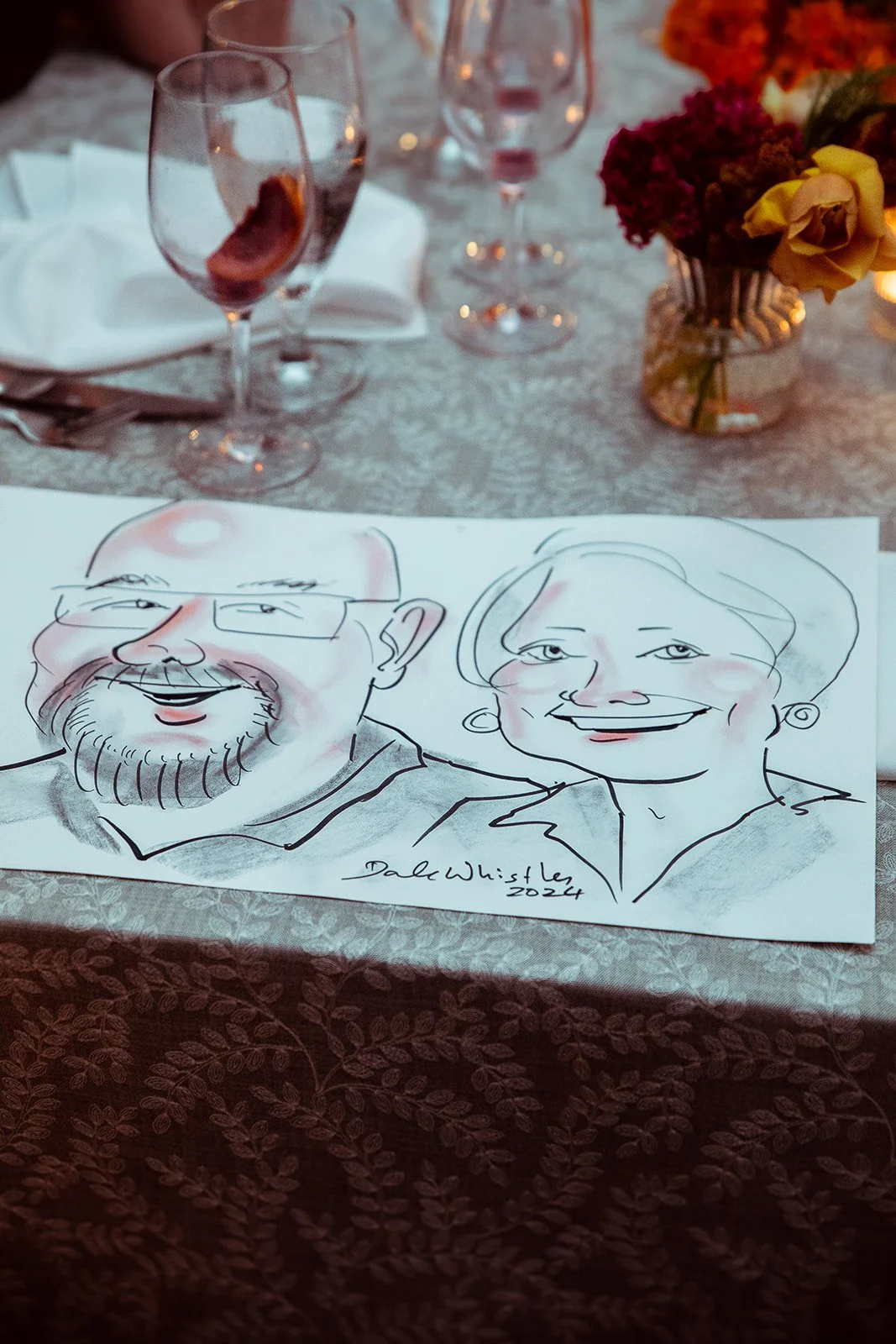 A sketch of two smiling people, a man with glasses and a beard, and a woman with short hair, is placed on a table set for a meal with wine glasses and a flower arrangement.