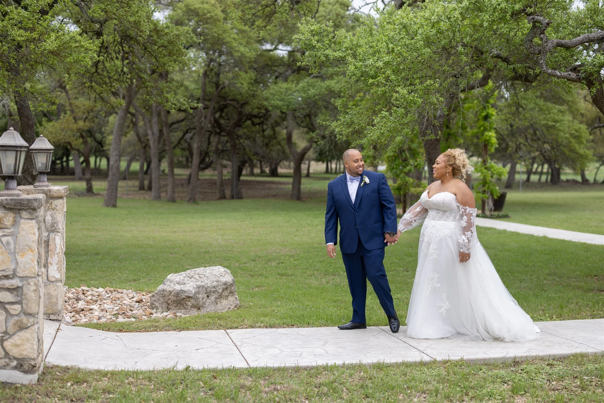 A newlywed couple holding hands and smiling at each other while walking outdoors on a paved pathway in a park or garden setting. The groom wears a dark blue suit with a bow tie and white boutonnière, and the bride wears a white strapless wedding dres