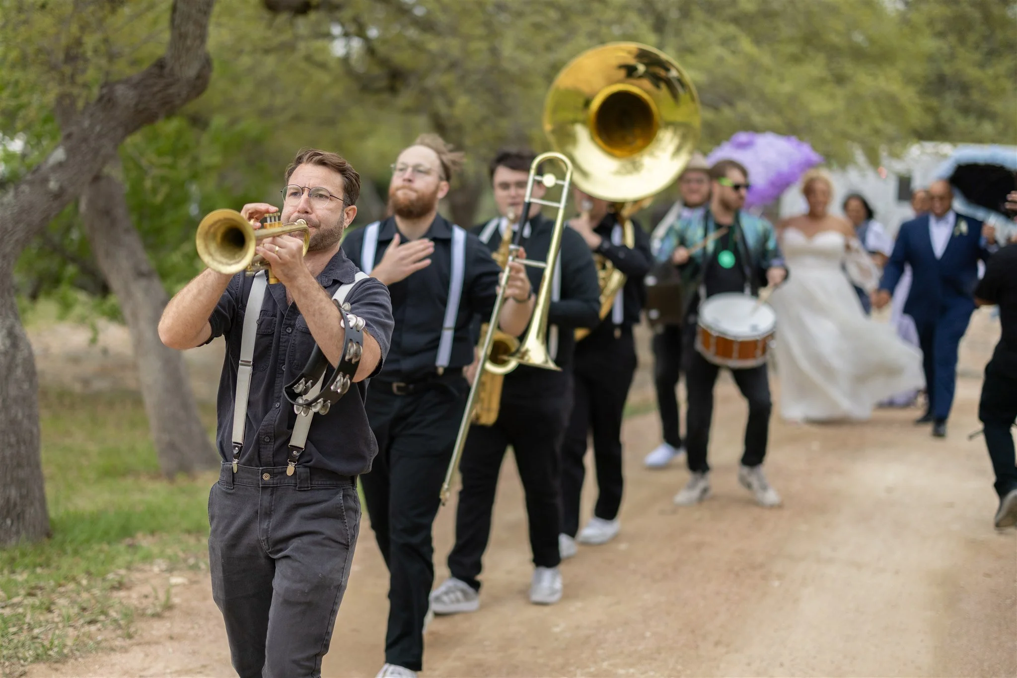 A band of musicians playing brass and percussion instruments in a procession, with a bride and groom in the background, outdoors on a dirt path with trees.