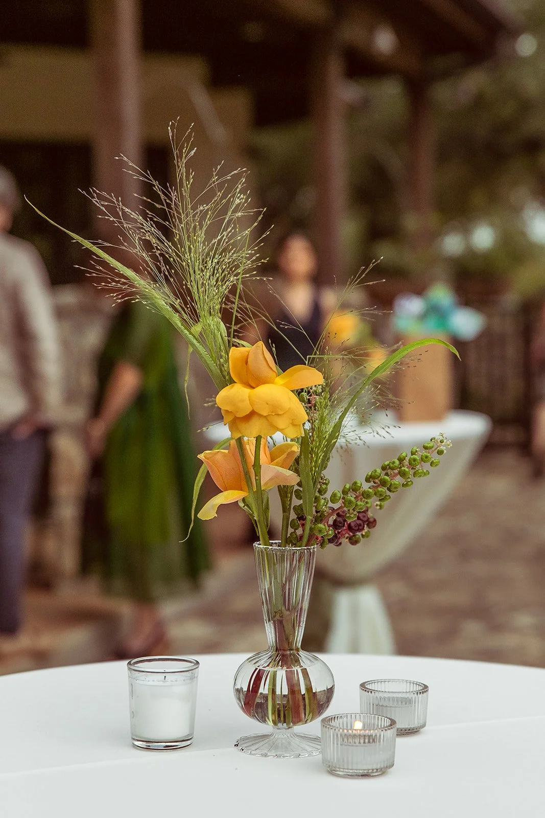 Flower arrangement in a glass vase on a white table, with a drinking glass and small candles nearby, in a blurred background of people and outdoor setting.