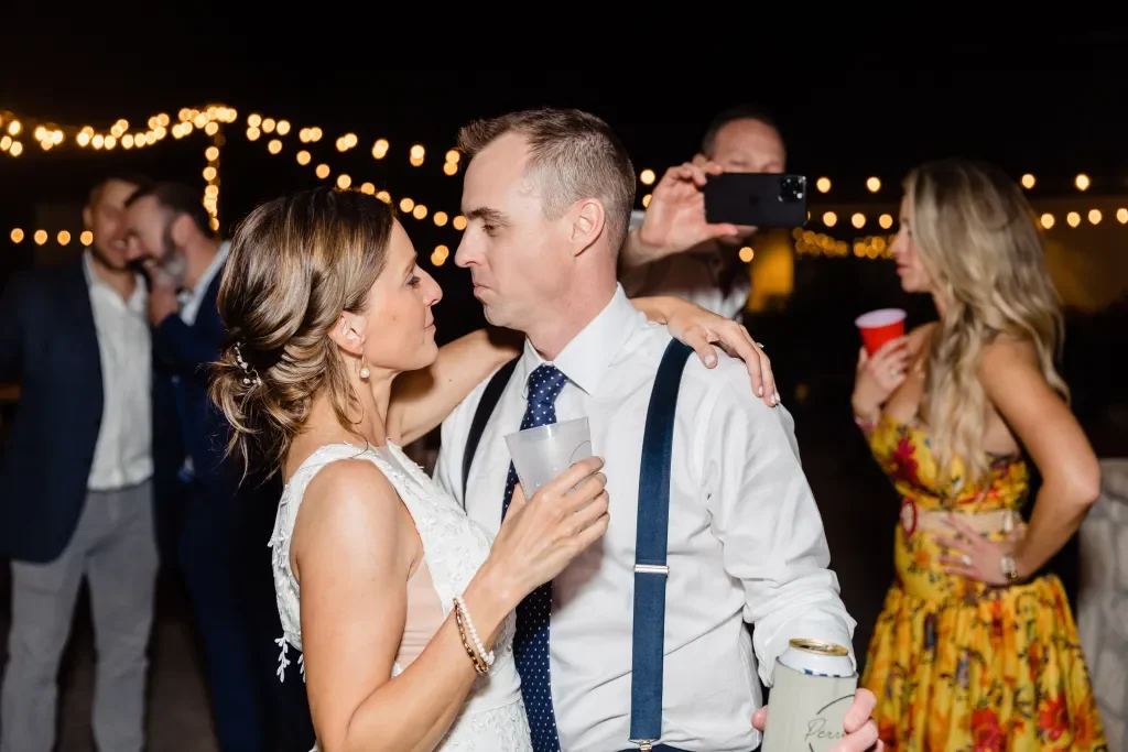 A couple dances closely at a night outdoor event with string lights, surrounded by other people with drinks and poses.