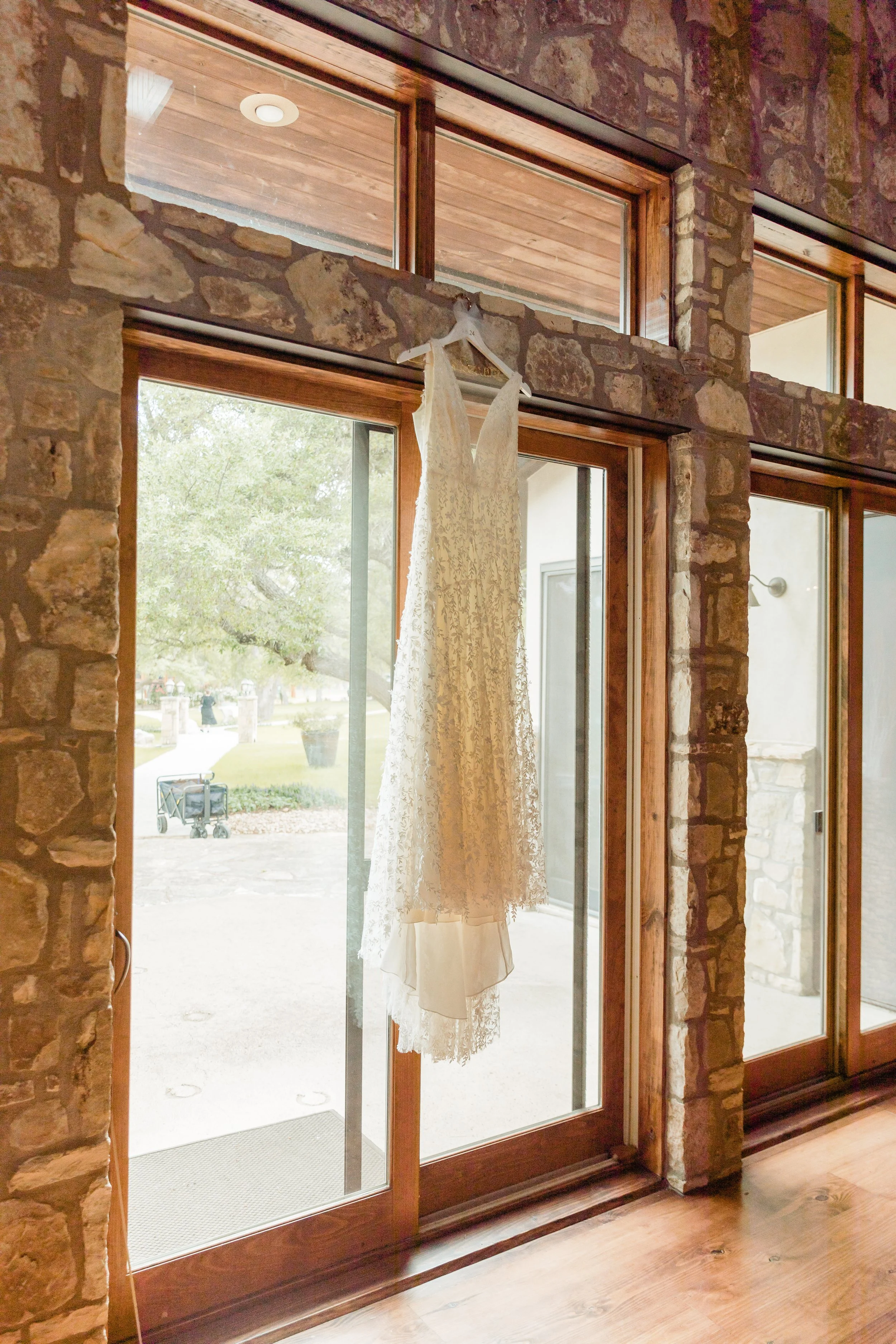 A white lace wedding dress hanging from a hanger inside a building with brick walls and large glass doors.