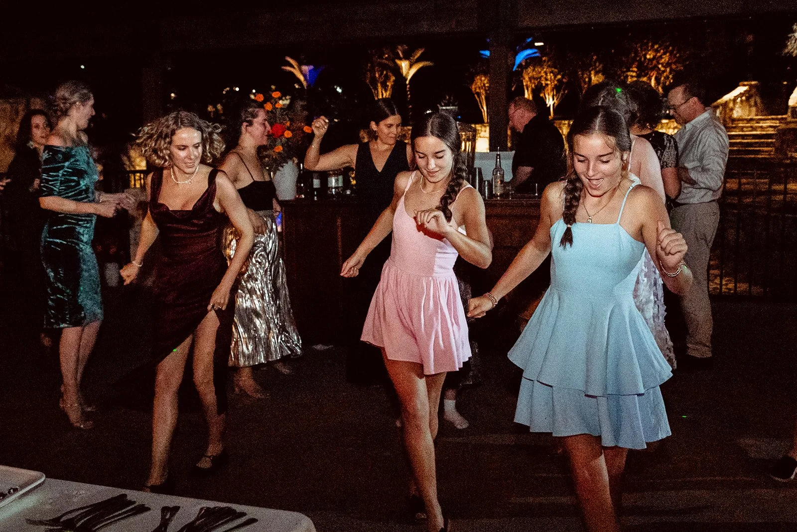 People dancing at a lively party or celebration, some women in colorful dresses, with a bar and tropical lighting in the background.