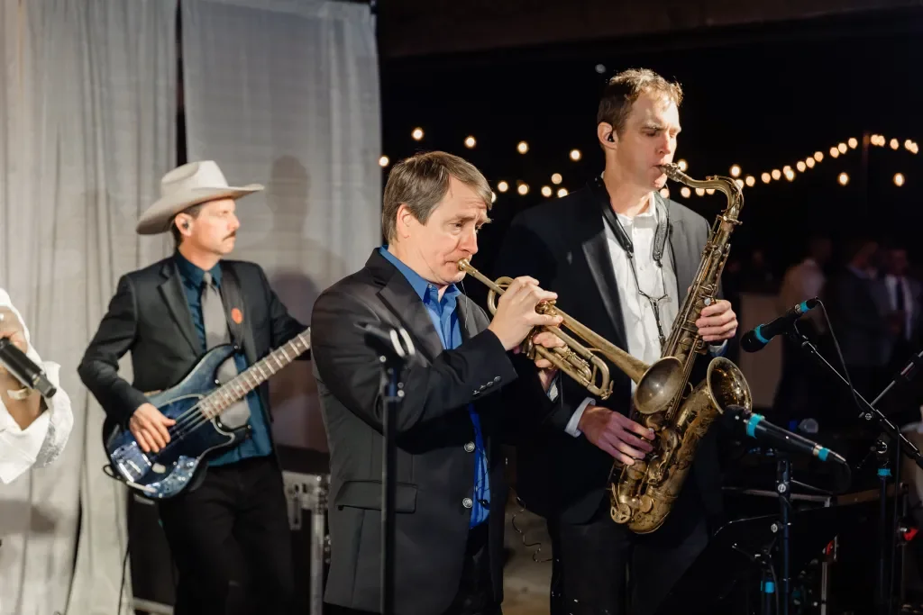 Three male musicians playing instruments at a live event: one on electric guitar, one on trumpet, and one on saxophone, with string lights in the background.