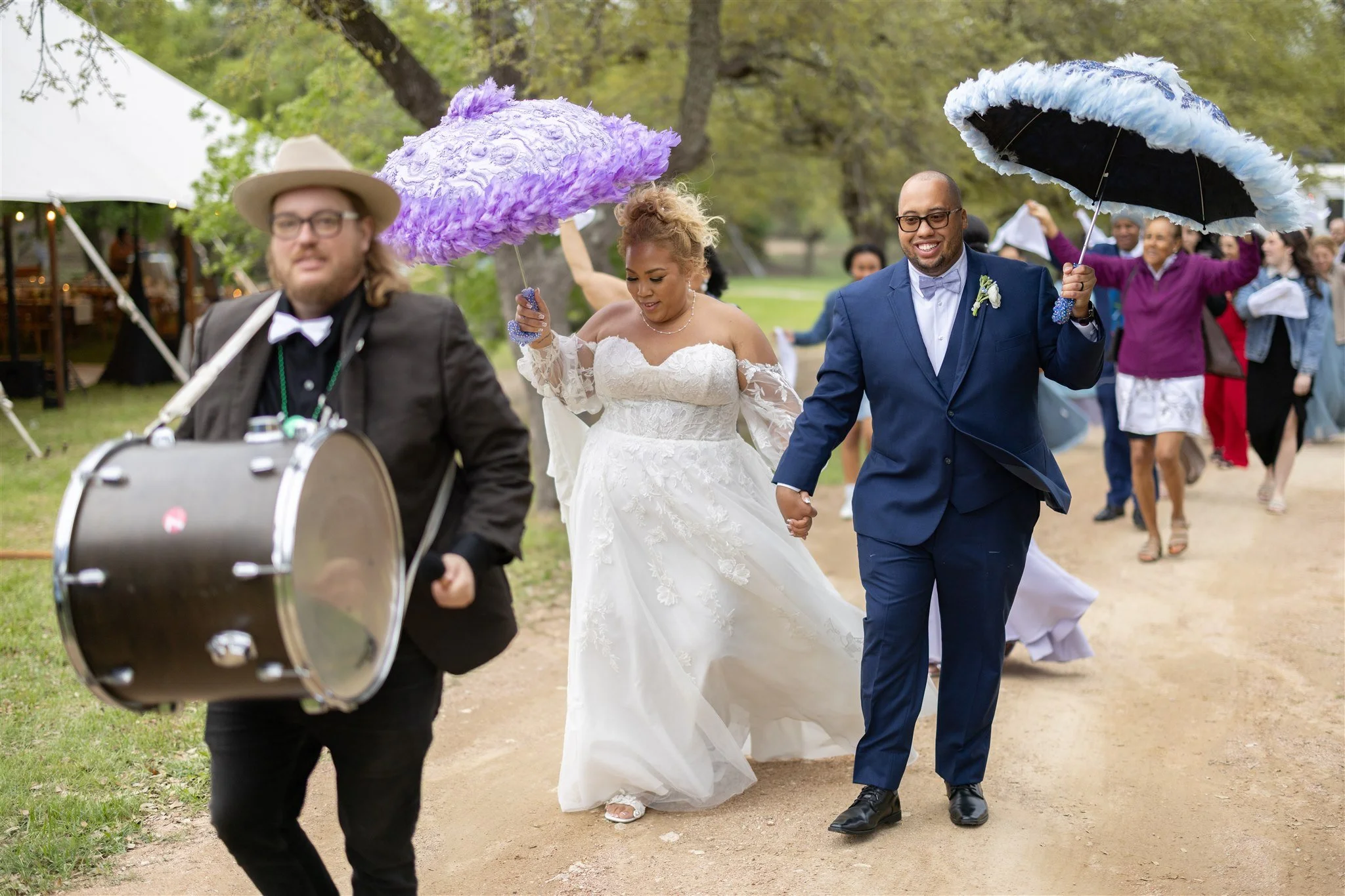 A wedding procession outdoors, with a bride and groom holding hands and smiling, walking on a dirt path surrounded by guests holding umbrellas, and a musician playing drums leading the way.