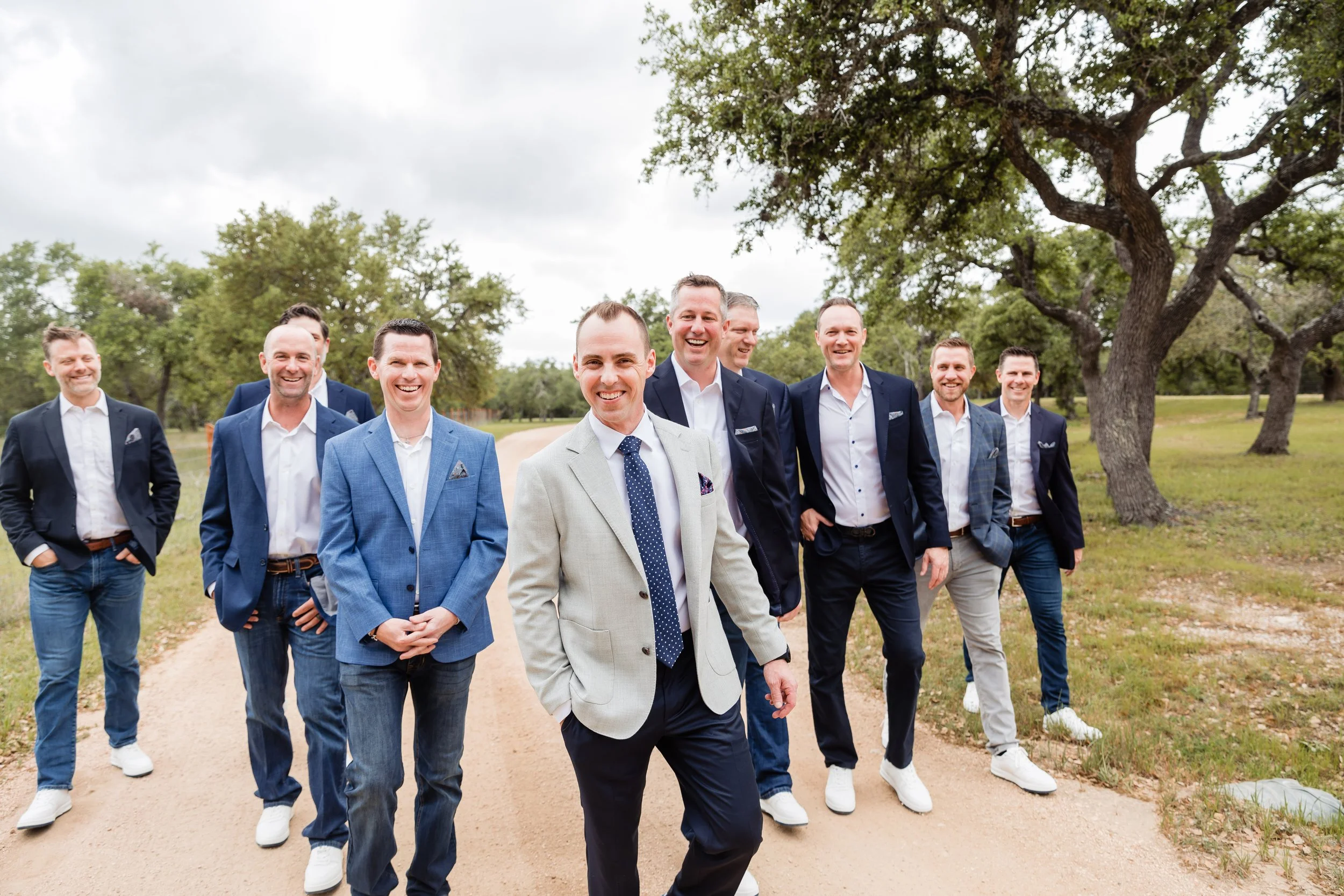 Group of men wearing suits and casual shoes walking outdoors on a dirt path with trees in the background, smiling and enjoying each other's company.