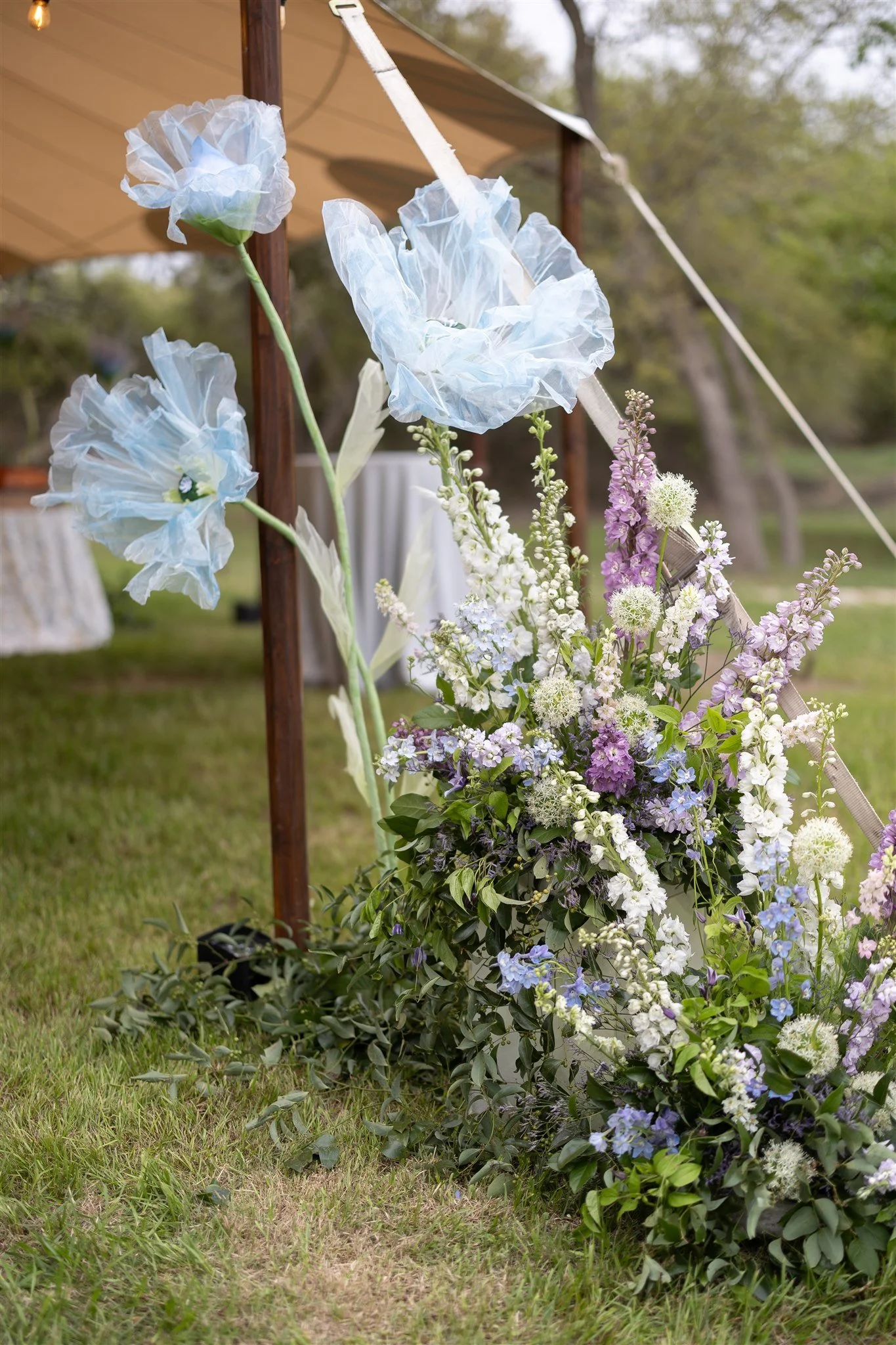 A floral arrangement with white and purple flowers, some resembling poppies, and green foliage, placed outdoors on the grass beside a brown wooden post. There are also large, white, tissue-paper-like flowers attached to the post, part of decorative e