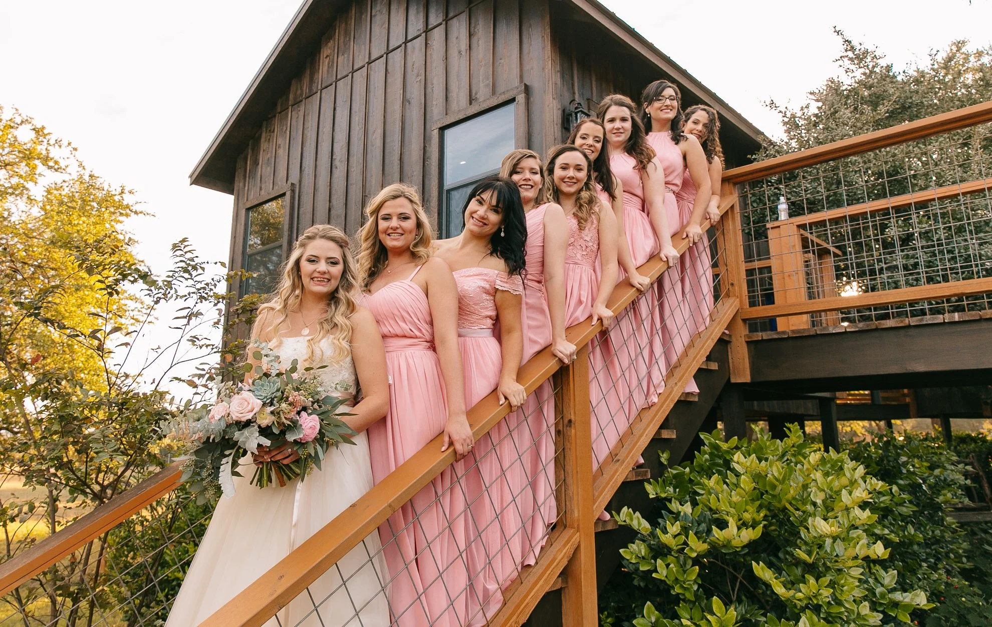 A bride and her bridesmaids standing on outdoor wooden staircase, smiling at camera, with the bride holding a bouquet, in front of a rustic wooden house, surrounded by trees and bushes.