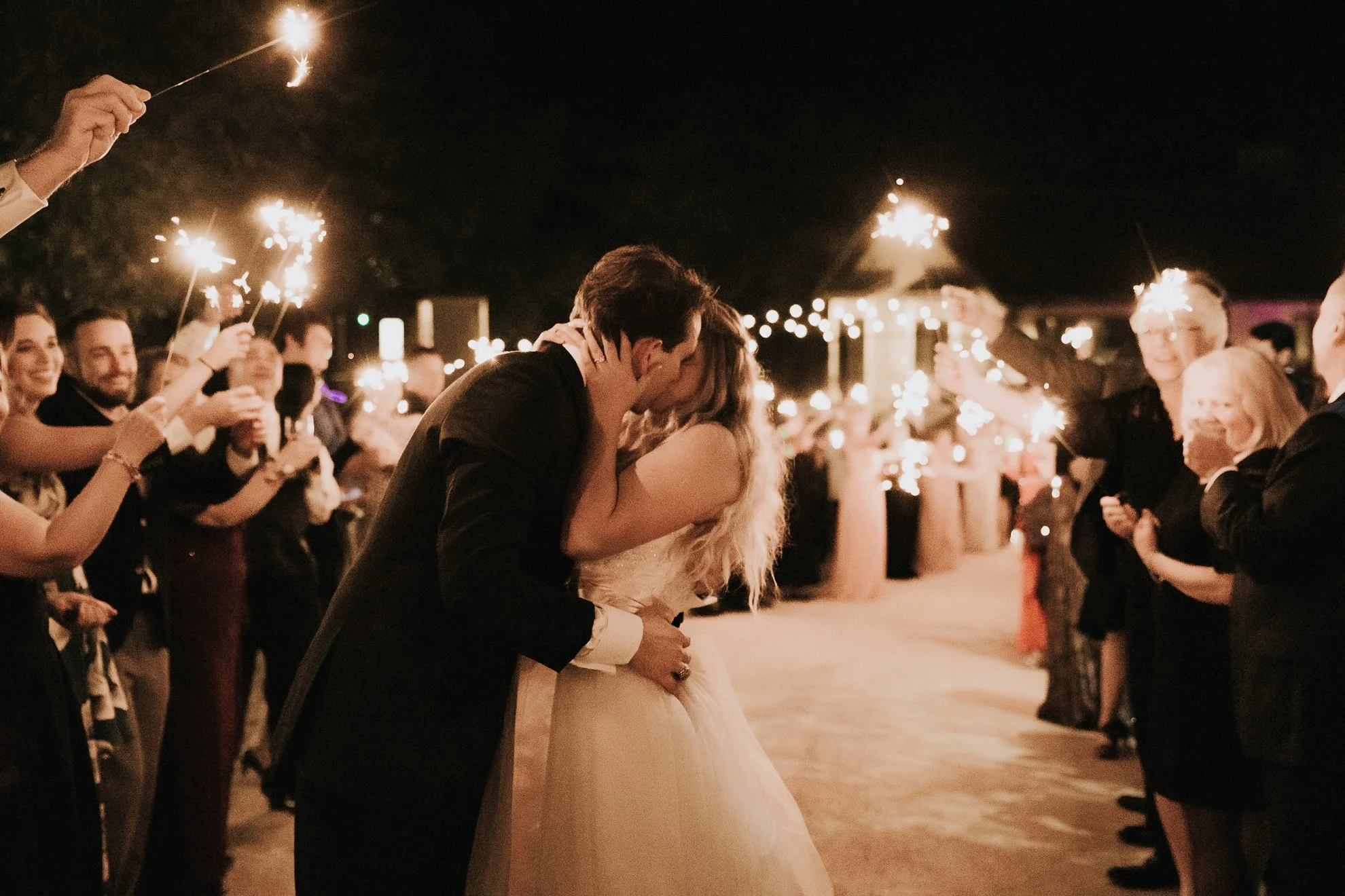 A newlywed couple sharing a kiss at night during their wedding celebration, surrounded by guests holding sparklers.
