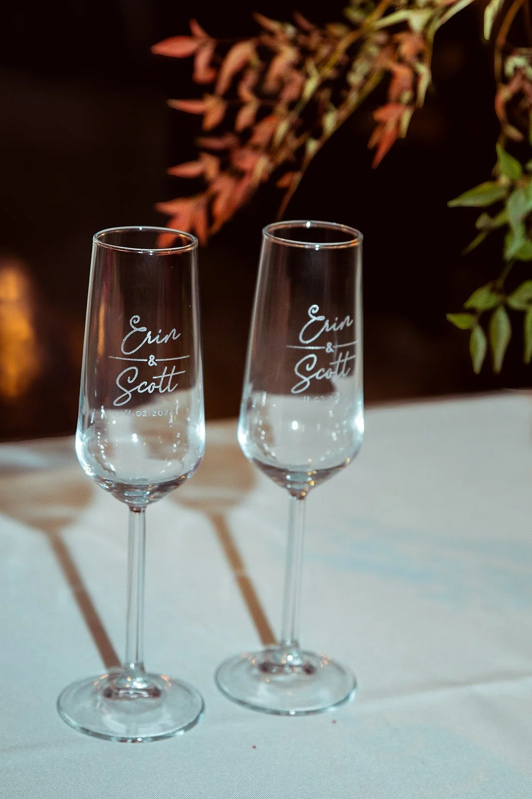 Two empty champagne flutes with engraved names Erin & Scott sit on a white table, with leaves in the background.
