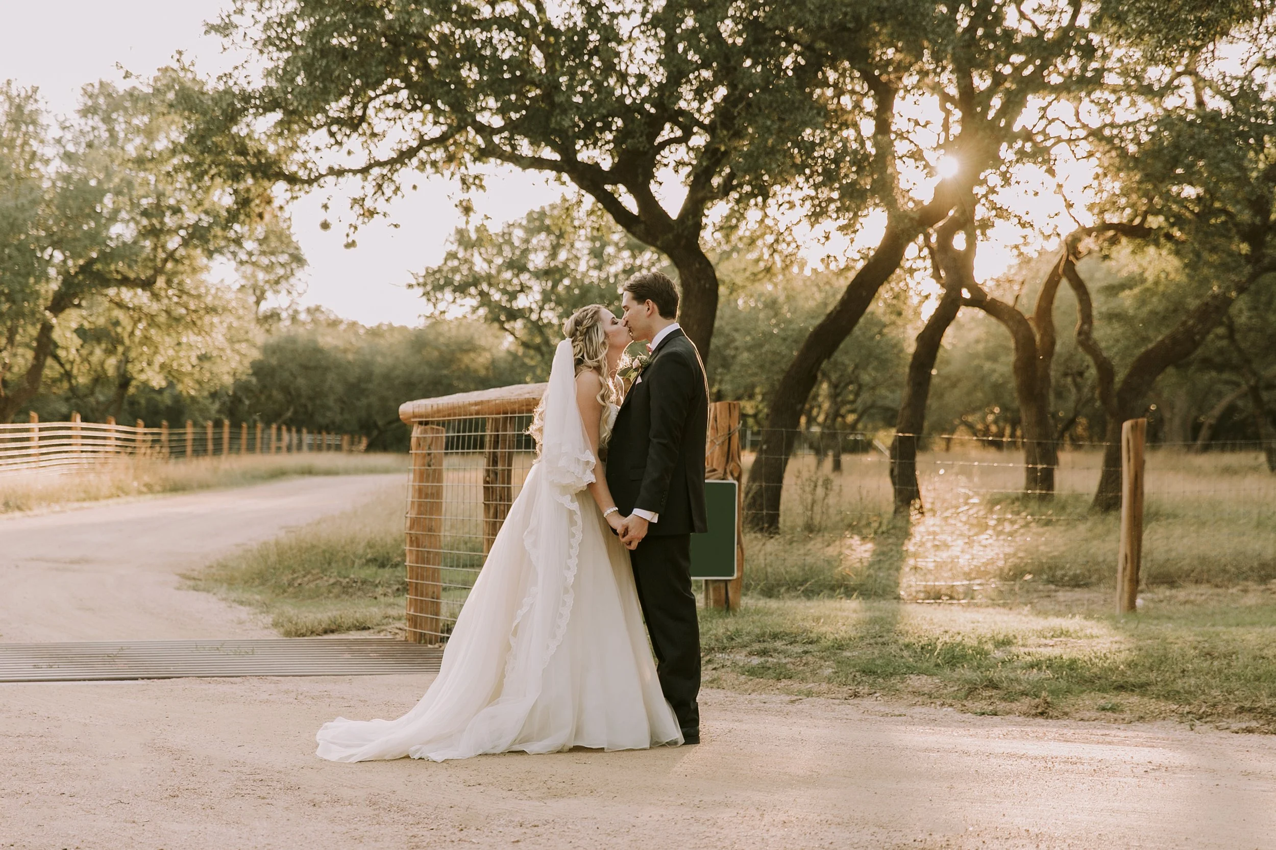 A bride and groom sharing a kiss in a rural outdoor setting during sunset, holding hands, with trees and a dirt path in the background.