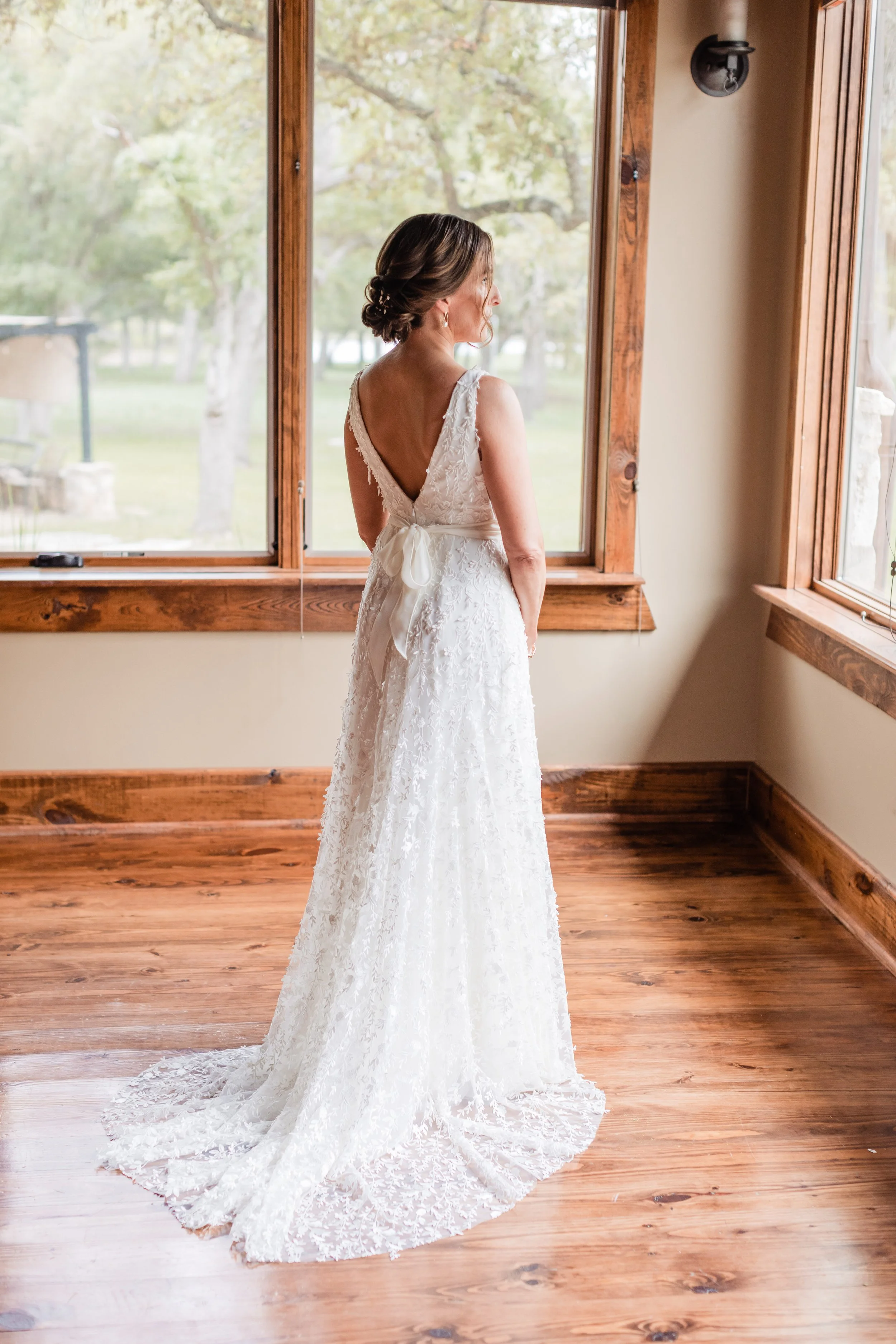 A bride standing indoors, wearing a white lace wedding gown with a low back and a bow tie, looking out the window at a green outdoor scene.