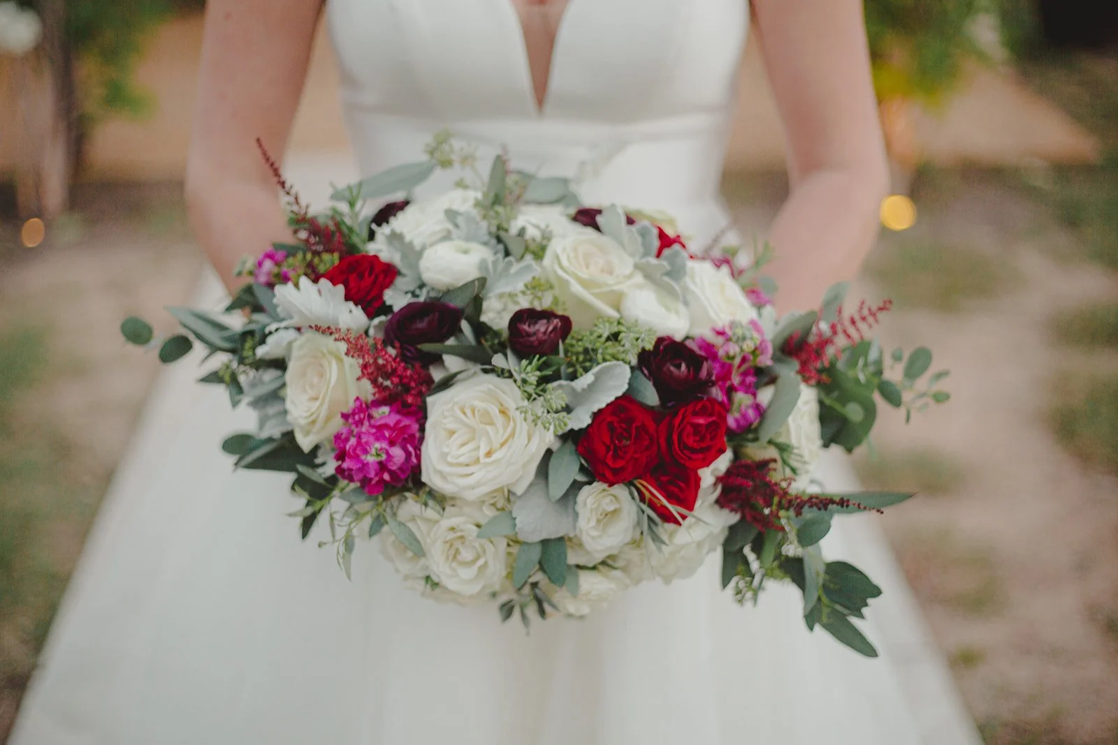A bride in a white wedding dress holding a bouquet of red, white, purple, and pink flowers with green foliage.