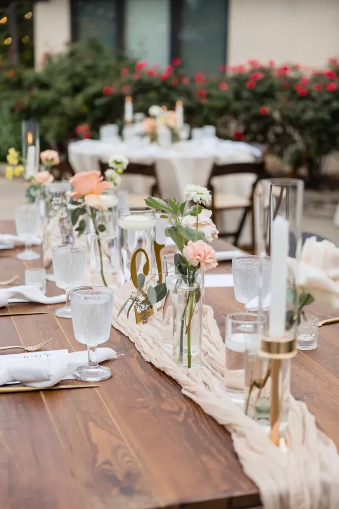 Outdoor wedding reception table decorated with pink and white flowers, candles, and glassware, with a backyard garden backdrop.