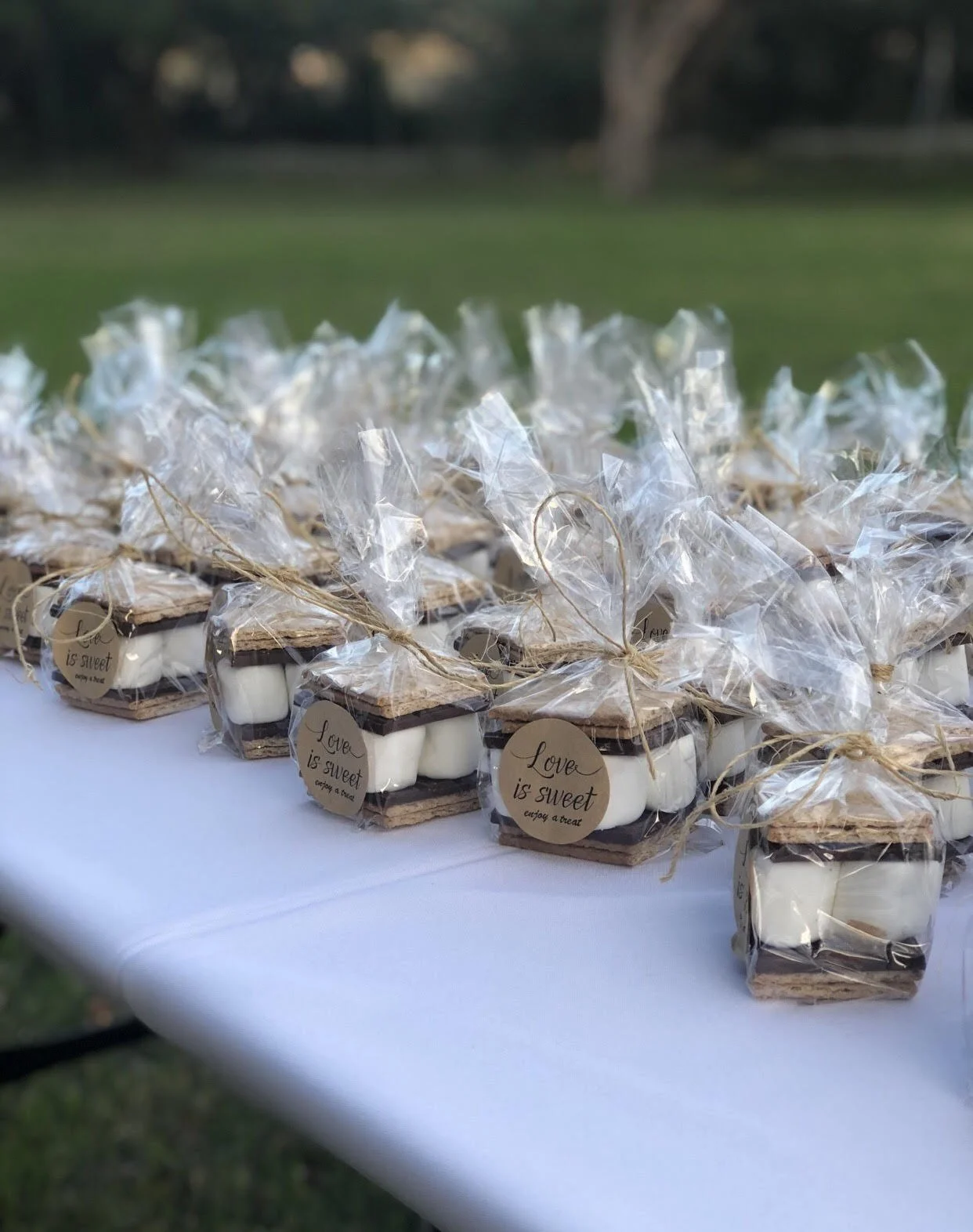 Small gift boxes containing chocolates, wrapped in clear plastic with a string and a round tag reading 'Love is sweet, enjoy a treat,' are arranged on a white table outdoors.