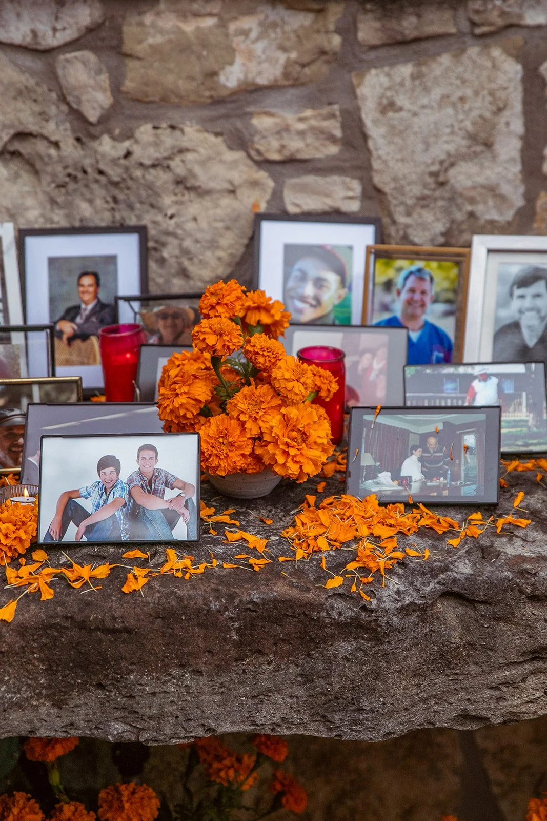 A memorial display featuring framed photographs of various people, an orange marigold flower arrangement, and scattered orange flower petals on a stone surface, set against a stone wall background.