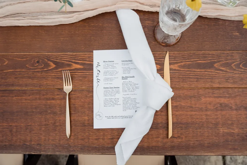 A wooden table setting with a menu card in the center, a gold fork on the left, and a gold knife on the right wrapped with a white cloth napkin. A glass of water with a yellow flower is partially visible at the top right.