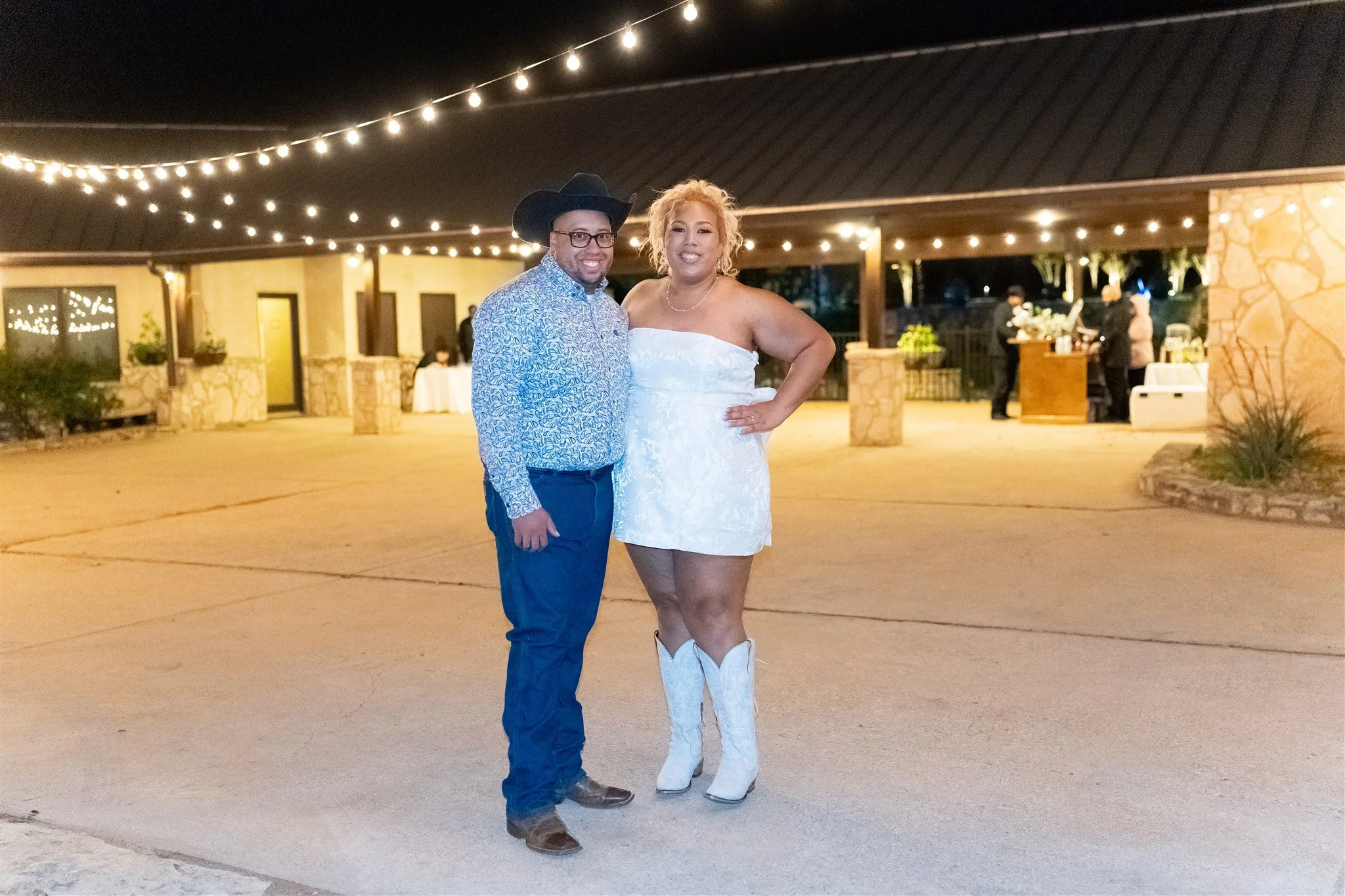A man in a patterned shirt and cowboy hat standing next to a woman in a white dress and cowboy boots, both smiling at night under string lights in an outdoor setting.