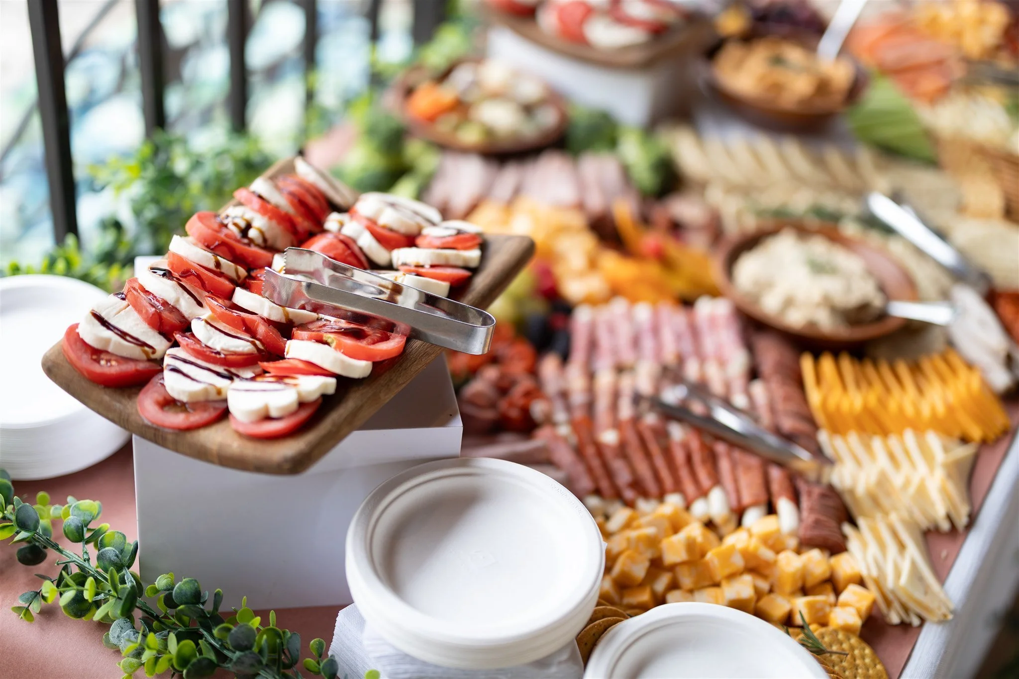 Assorted cold cuts, cheeses, and sliced vegetables on a buffet table with white plates, set outdoors.