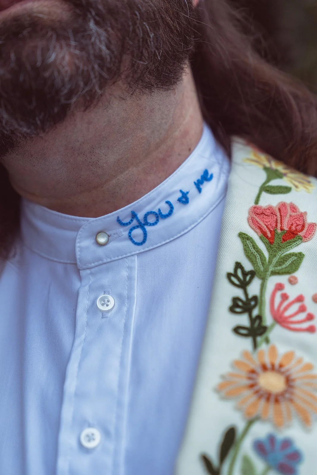 Close-up of a person wearing a white shirt with embroidered blue text that says 'You & Me' around the collar, and a cream-colored jacket with colorful embroidered flowers on the shoulder.
