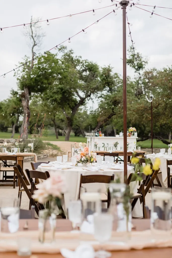 Outdoor wedding reception setup with round tables decorated with floral centerpieces, surrounded by wooden chairs, under string lights, with a white buffet table in the background, set in a lush green park.