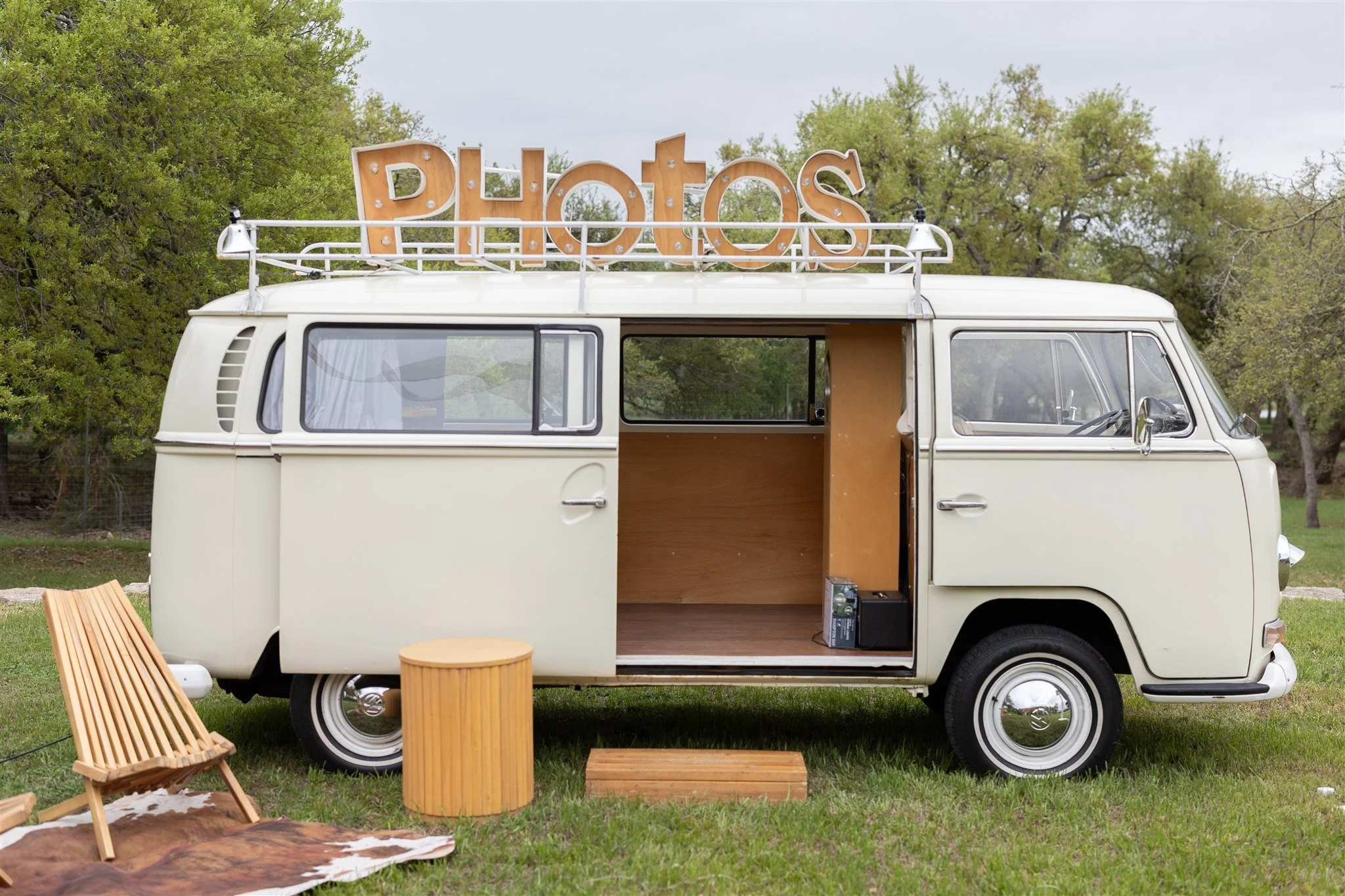 Vintage cream-colored van with open sliding door, parked on grass with trees in the background, a wooden bench, a cylindrical wooden side table, and a small wooden step in front. Large illuminated sign spelling 'PHOTOS' is mounted on the roof.