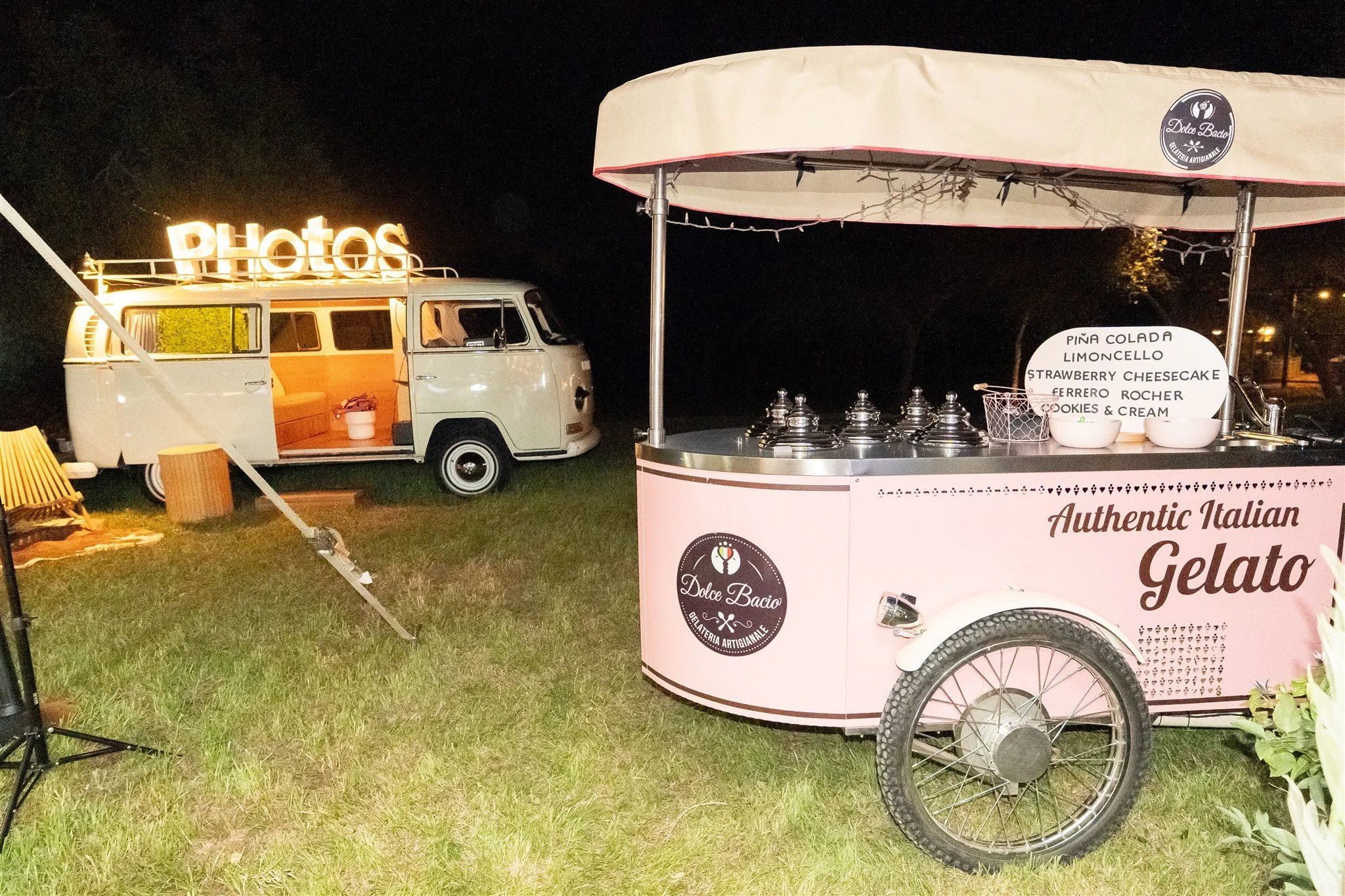 Pink gelato cart with the logo 'Dolce Bacio' and a white vintage van with a lit sign saying 'Photos' in the background, set up outdoors at night on a grassy area.