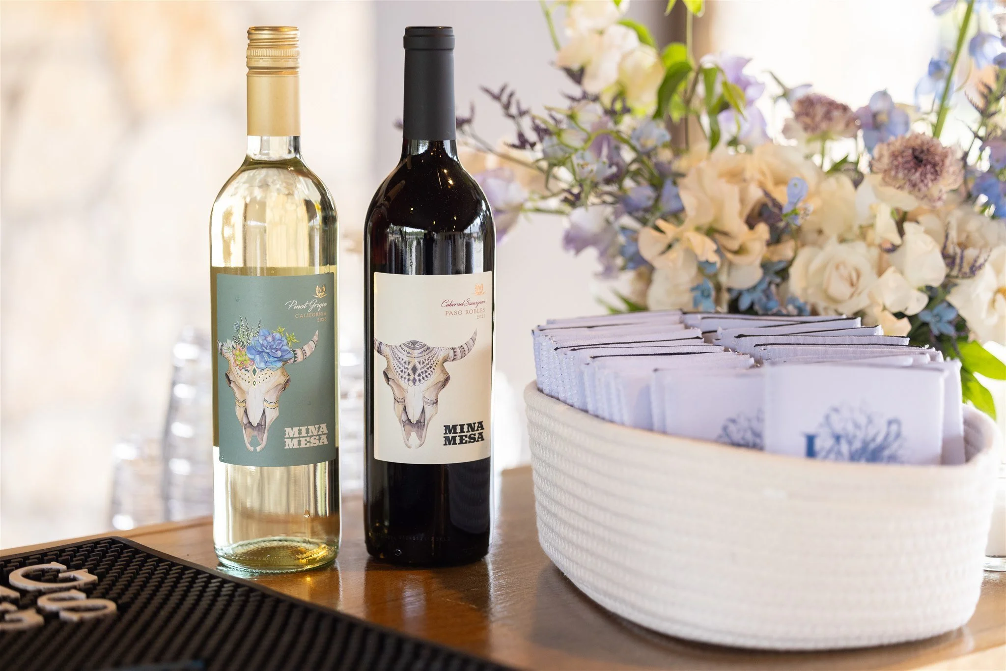 Two bottles of wine with animal skull labels, one white and one red, positioned on a wooden surface near a white woven basket containing napkins, with a bouquet of white, purple, and blue flowers in the background.
