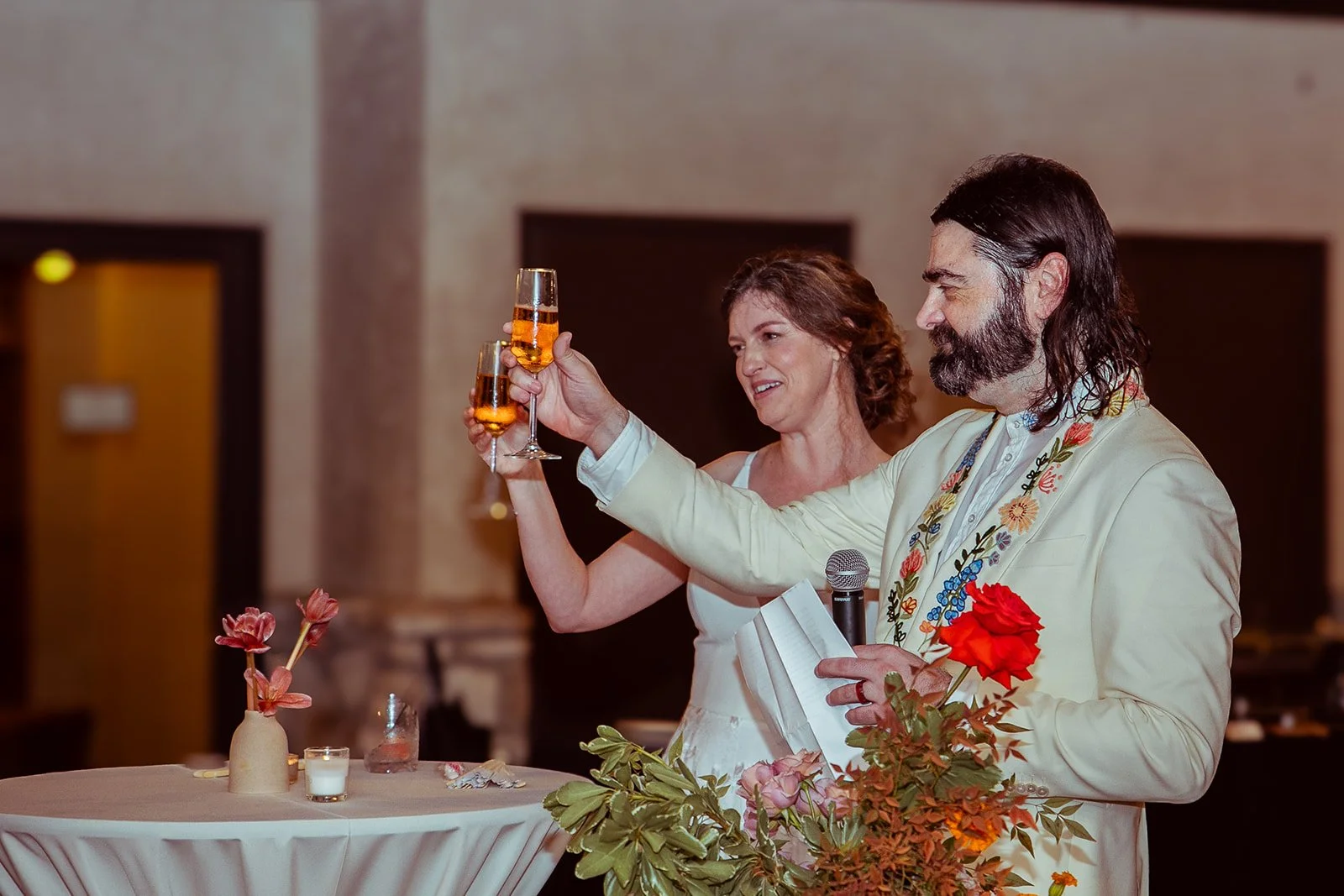 A man with long dark hair and a beard wearing a light-colored jacket with floral embroidery raises a champagne flute in a toast, standing next to a smiling woman with short curly hair, dressed in white, holding a champagne flute. They are at a table
