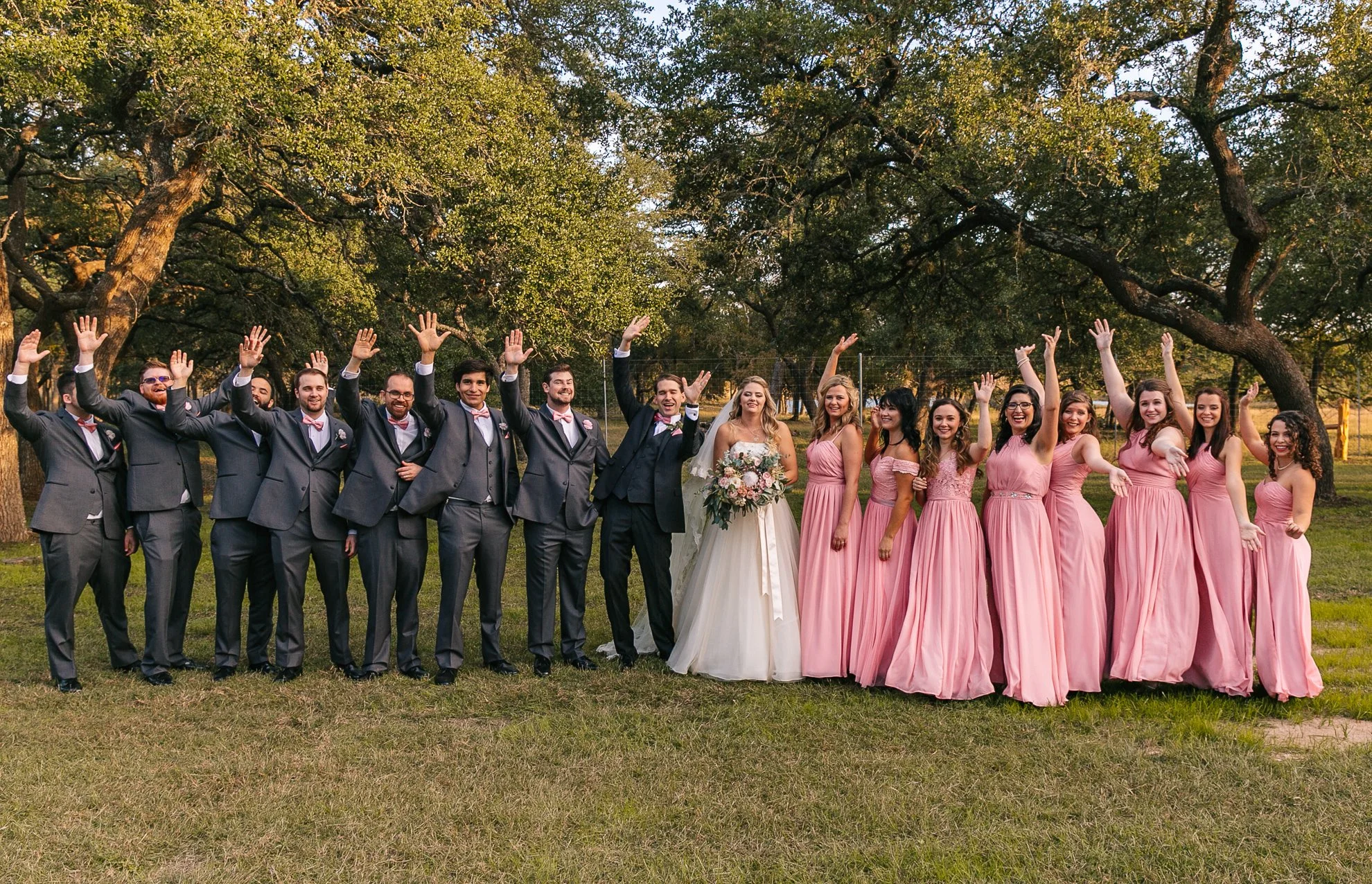 A wedding party with groomsmen in gray suits and bridesmaids in pink dresses stands outdoors on grass, raising their hands, in front of trees, during sunset.