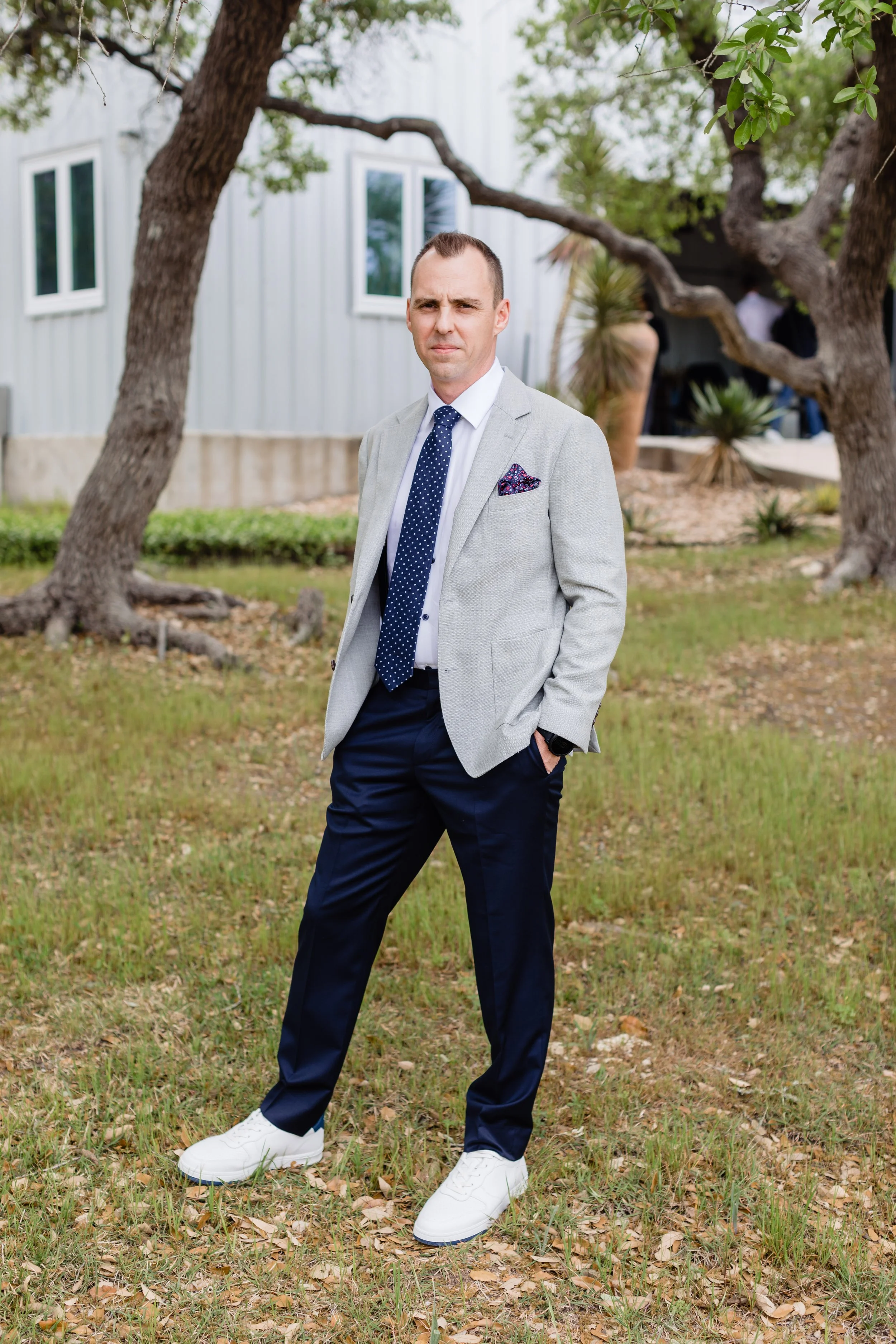 A man in a light gray blazer, white shirt, navy pants, and white sneakers standing outdoors with hands in pockets, under trees with a building in the background.