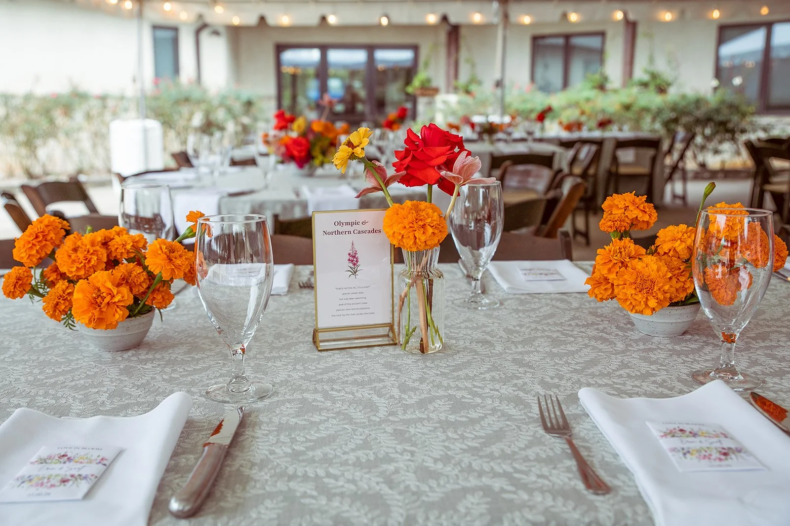 Elegant dining table decorated with orange marigold and red flowers in vases, set with white napkins, glassware, and silverware, in a restaurant or event space.